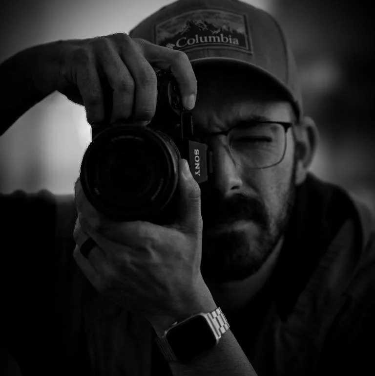 A man wearing glasses, a Columbia cap, and a smartwatch taking a black and white photo with a Sony camera.