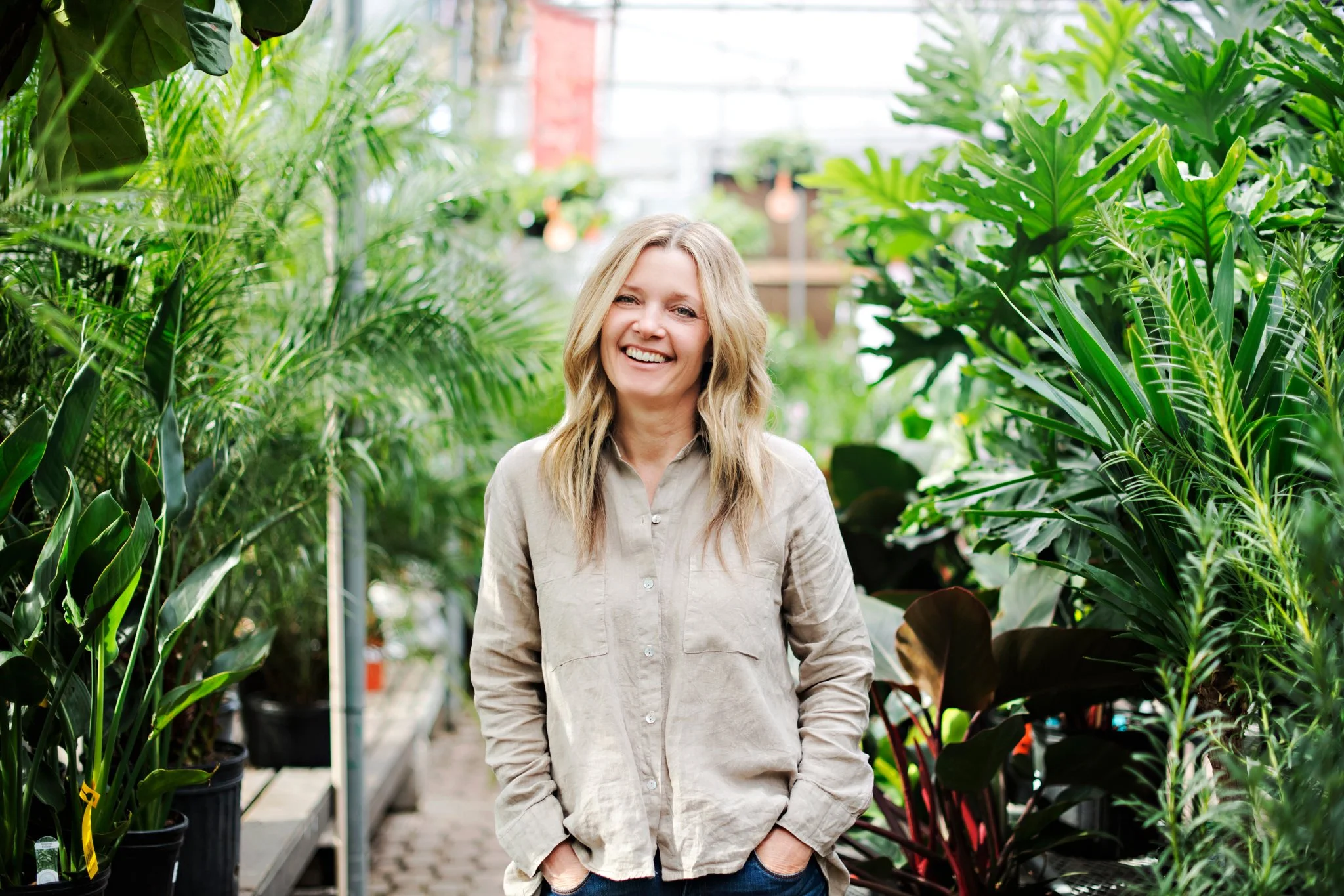 A smiling woman with blonde hair wearing a beige shirt standing amidst green plants in a greenhouse.