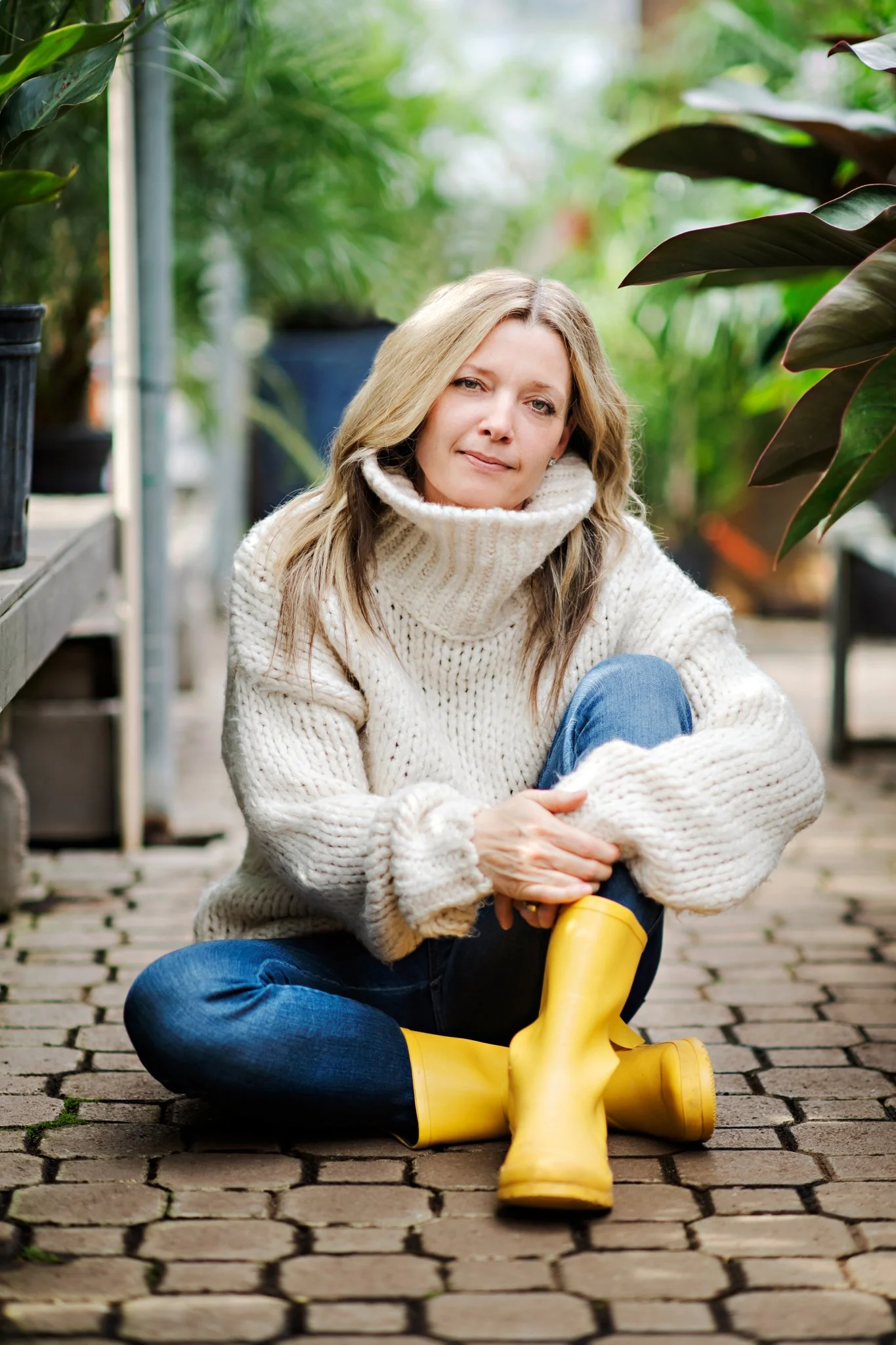 A woman sitting on a cobblestone path in a greenhouse, wearing a thick cream sweater, blue jeans, and yellow rain boots, surrounded by green plants.