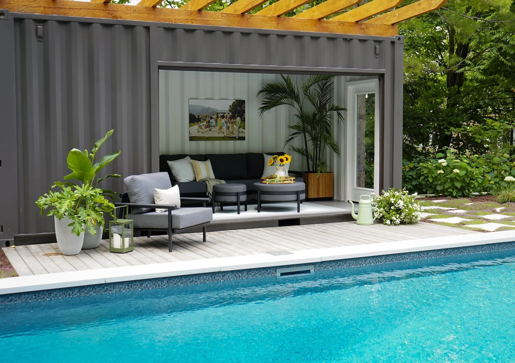 Outdoor pool area with a modern gray container home and a covered seating area. The seating area includes a black sectional sofa with white and yellow pillows, and two round ottomans. There is a small table with a sunflower arrangement, and potted pl