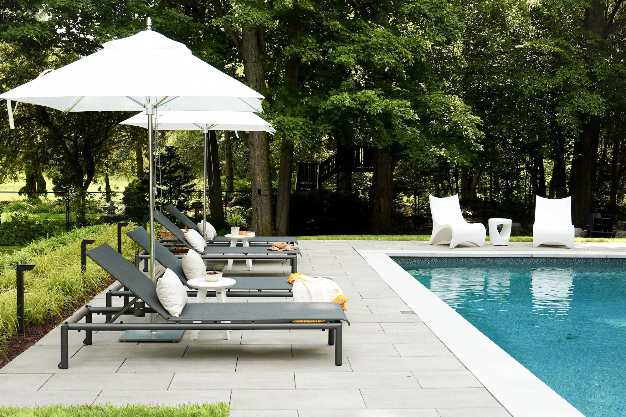 Outdoor swimming pool with lounge chairs, umbrellas, and modern white chairs surrounded by trees and greenery