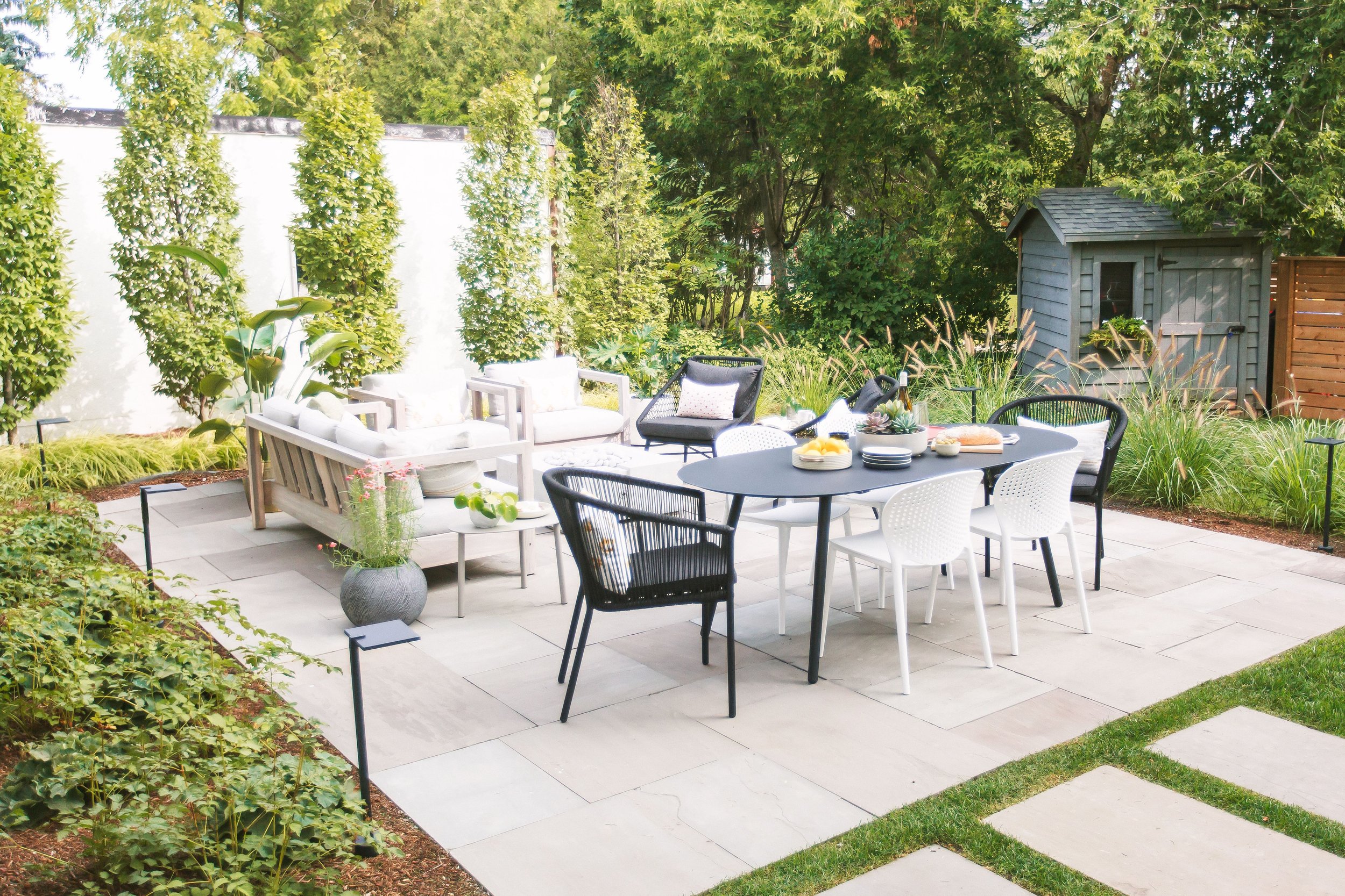 Outdoor patio with a black and white dining table, assorted chairs, and seating area surrounded by lush green plants and trees, with a small shed in the background.