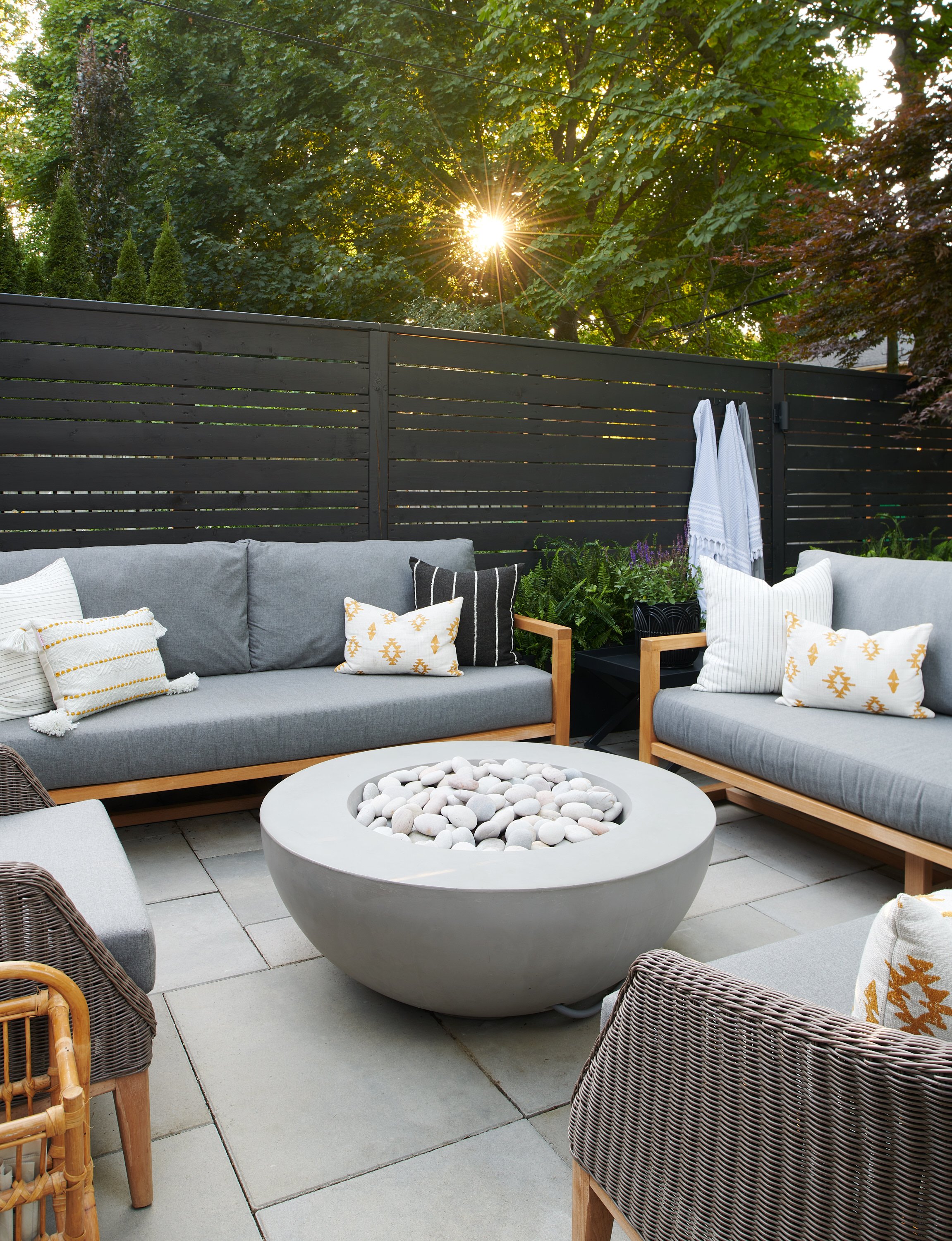 Outdoor patio with gray sofas, wicker chairs, and a round fire pit filled with white stones. Sun setting behind trees and a black fence in the background.