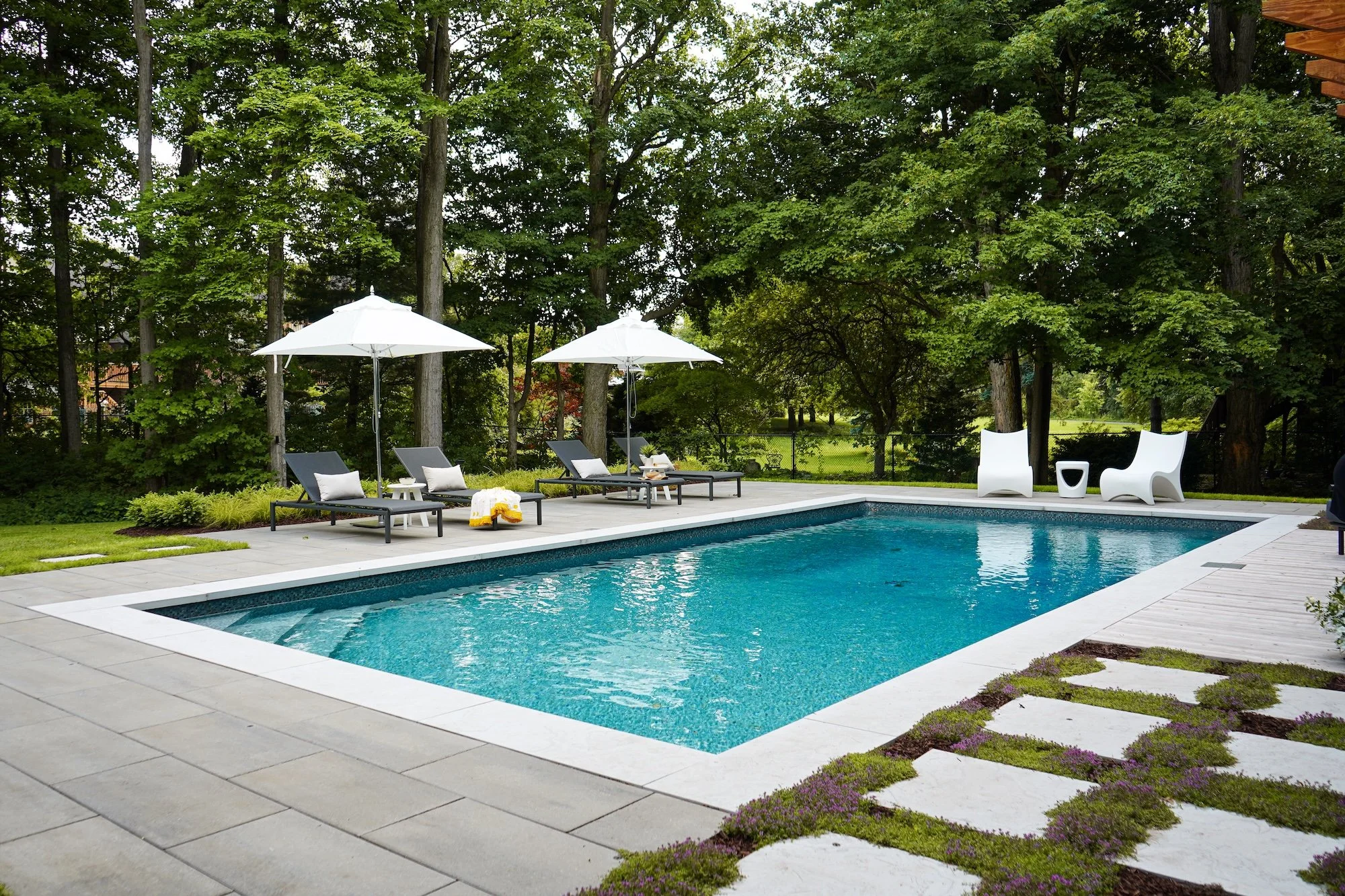 A backyard with a rectangular swimming pool surrounded by loungers with umbrellas and trees in the background.