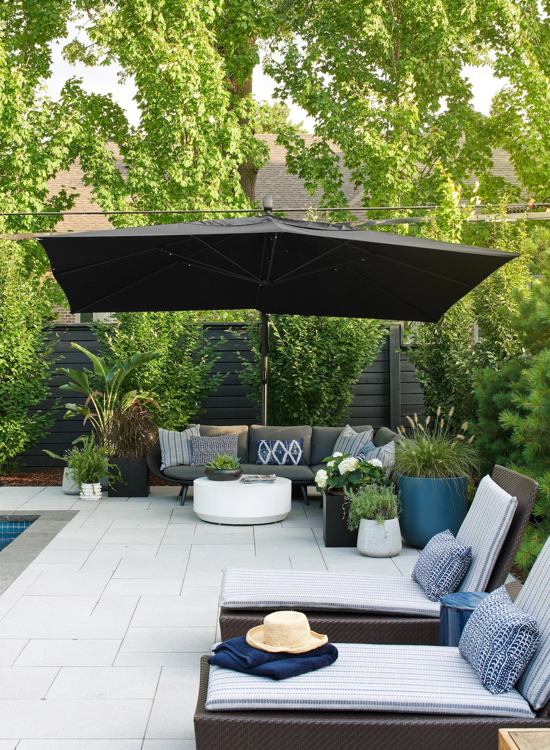 Outdoor patio area with two lounge chairs, a sectional sofa, and a black umbrella providing shade. There are various potted plants, throwing pillows on the furniture, and a straw hat with a towel on one of the lounge chairs. Green trees and a dark fence surround the area.