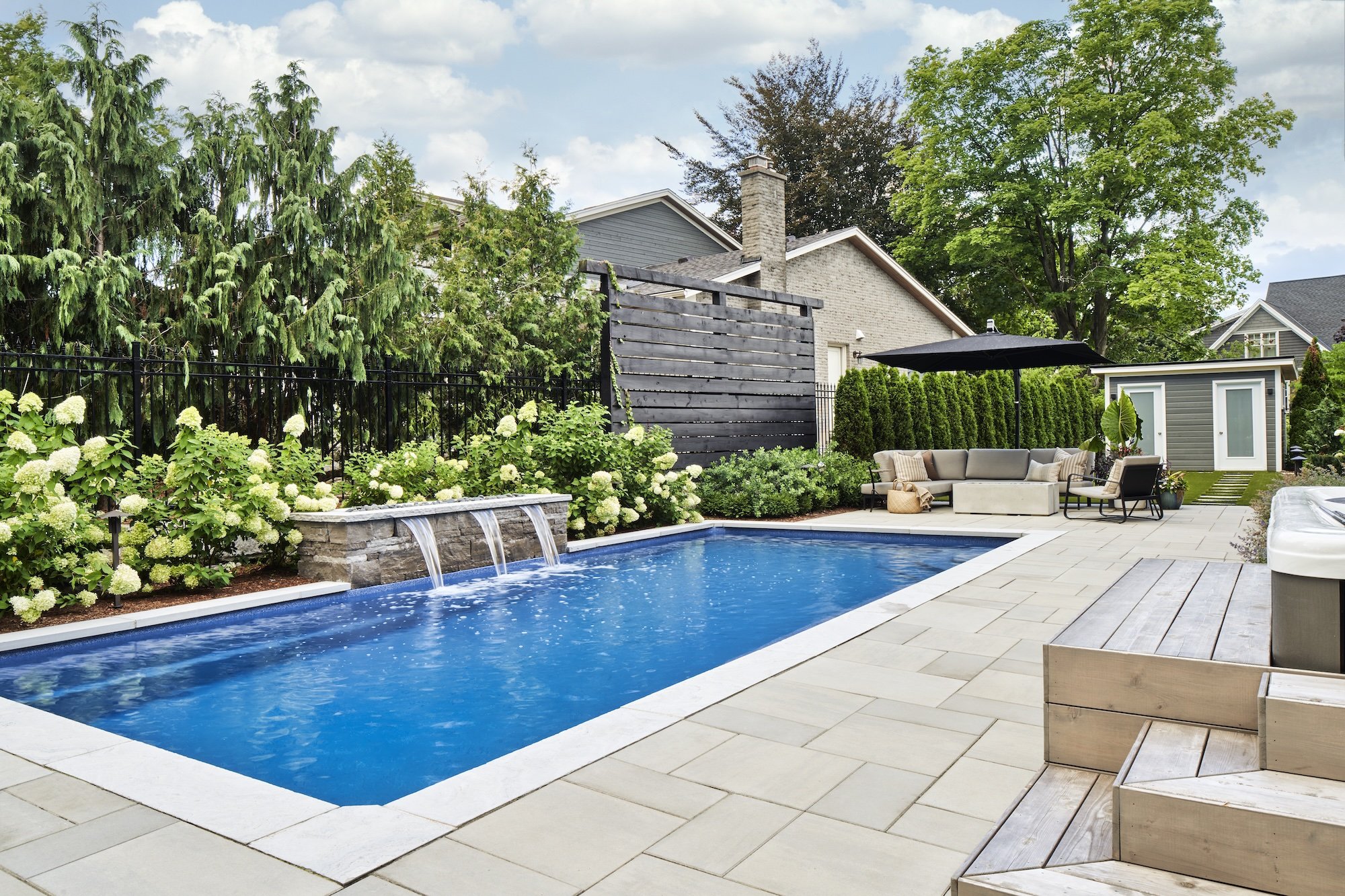 Backyard with a rectangular swimming pool, surrounded by a paved deck, lush green trees, shrubs, and white flowers, with outdoor seating including a sectional sofa under a black umbrella, and a small shed in the background.
