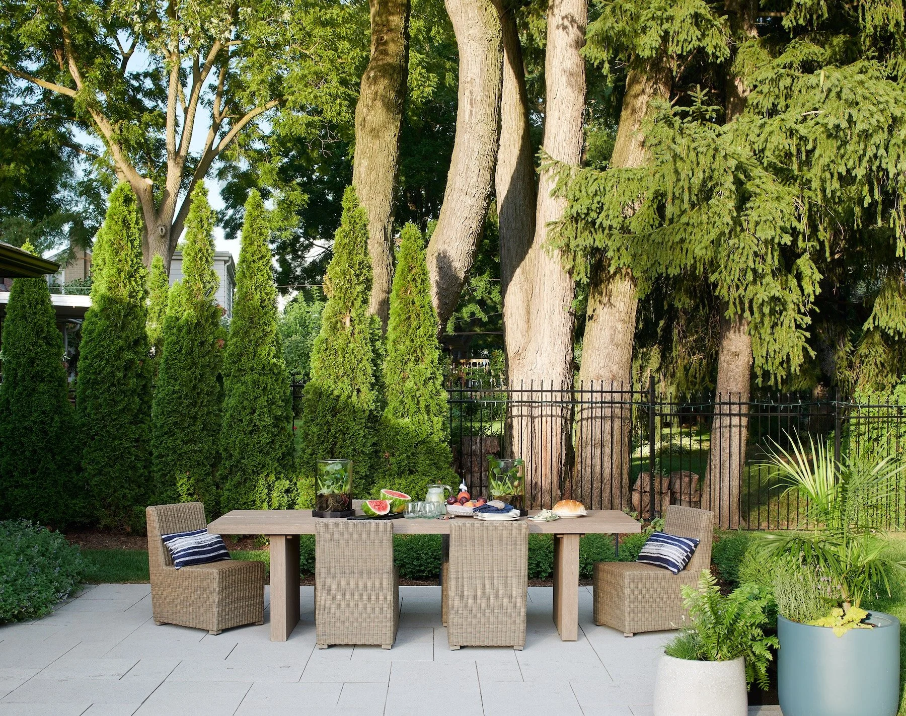 Outdoor patio dining table with chairs, fruits, bread, and drinks, surrounded by lush green trees and potted plants.