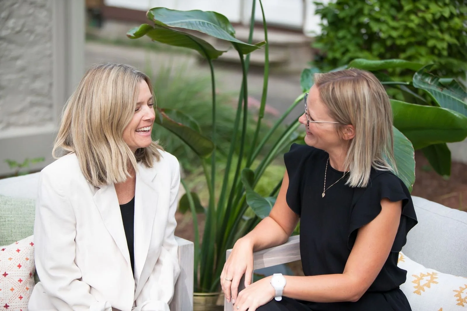 Two women are sitting outside, engaged in a cheerful conversation. The woman on the left has blonde hair and is wearing a white blazer, while the woman on the right has blonde hair, wears glasses, a black dress, and a watch. They are smiling and appear happy. There are large green plants and foliage in the background.
