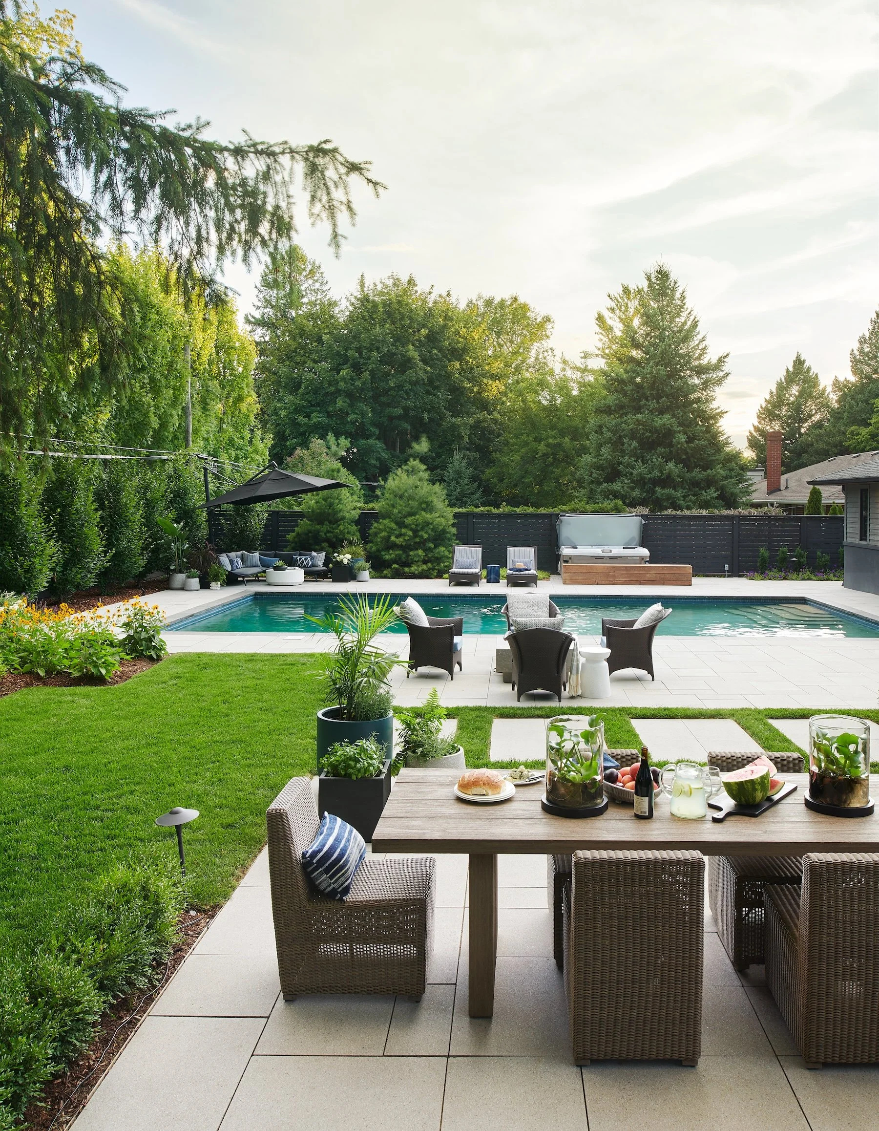 Backyard with swimming pool, outdoor furniture, potted plants, and a grassy lawn area, surrounded by trees and a fence.