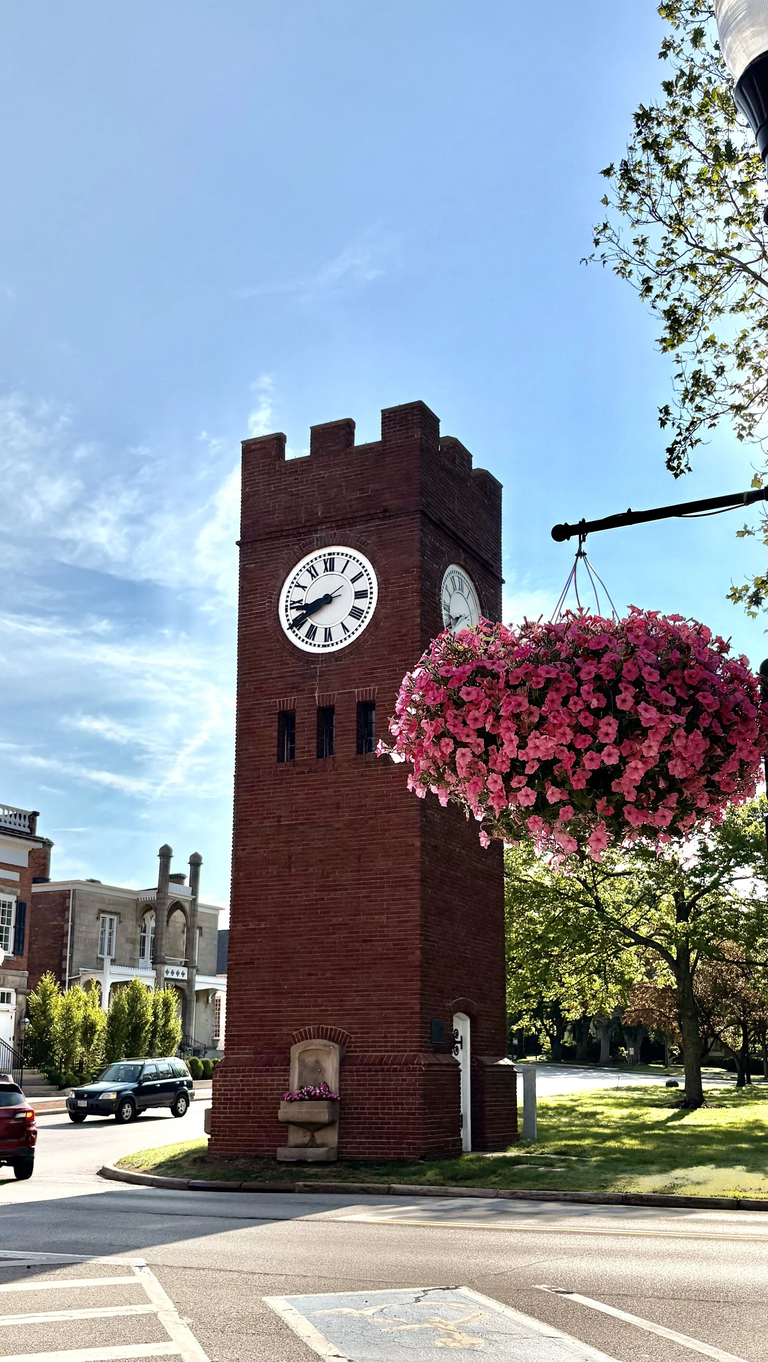 A tall red brick clock tower with Roman numerals on its face is situated in a town square, with a blue sky overhead and a hanging basket of pink flowers in the foreground.
