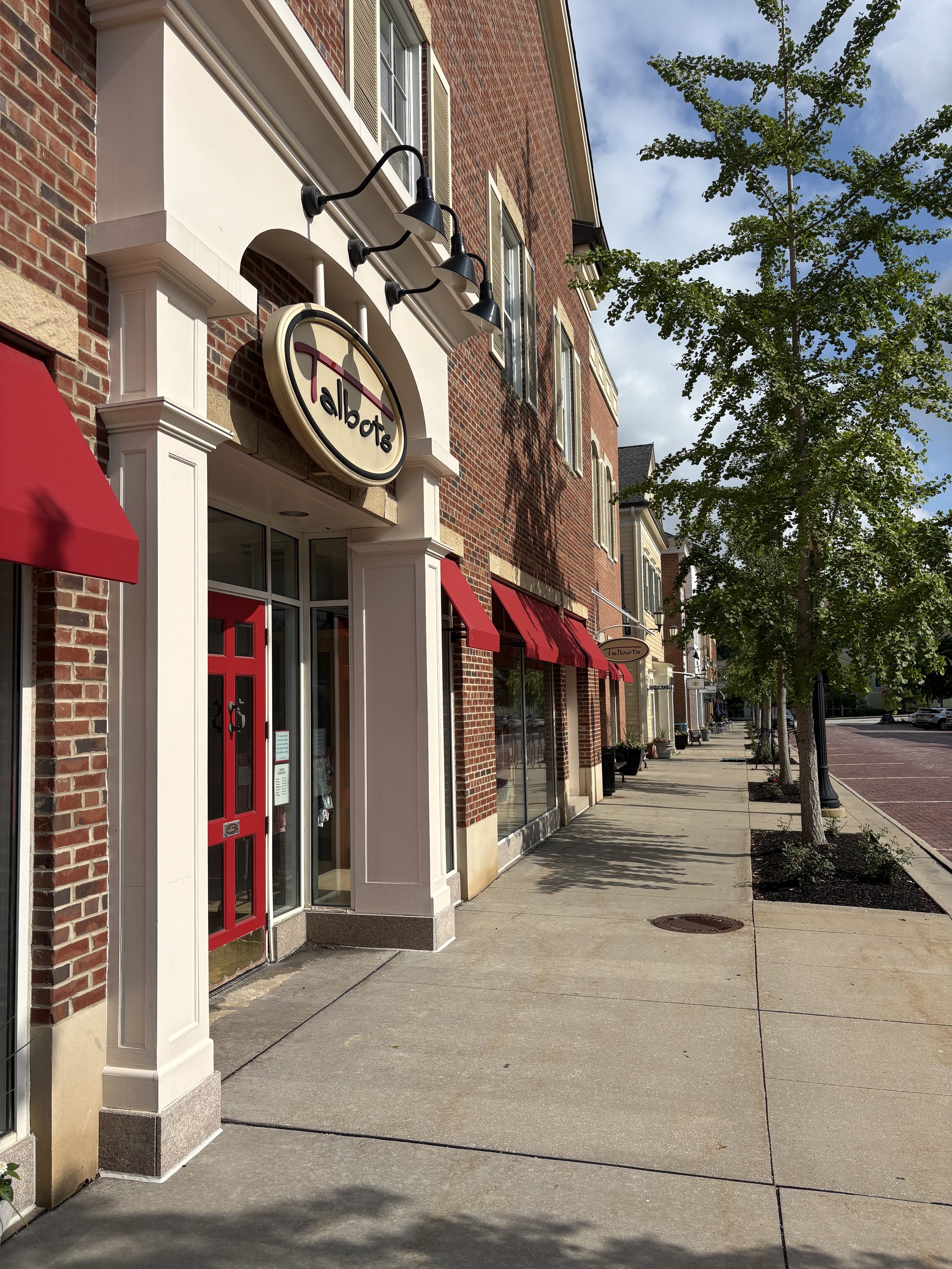 A sidewalk in front of a row of brick storefronts with red awnings, trees planted along the street, and a partly cloudy sky.