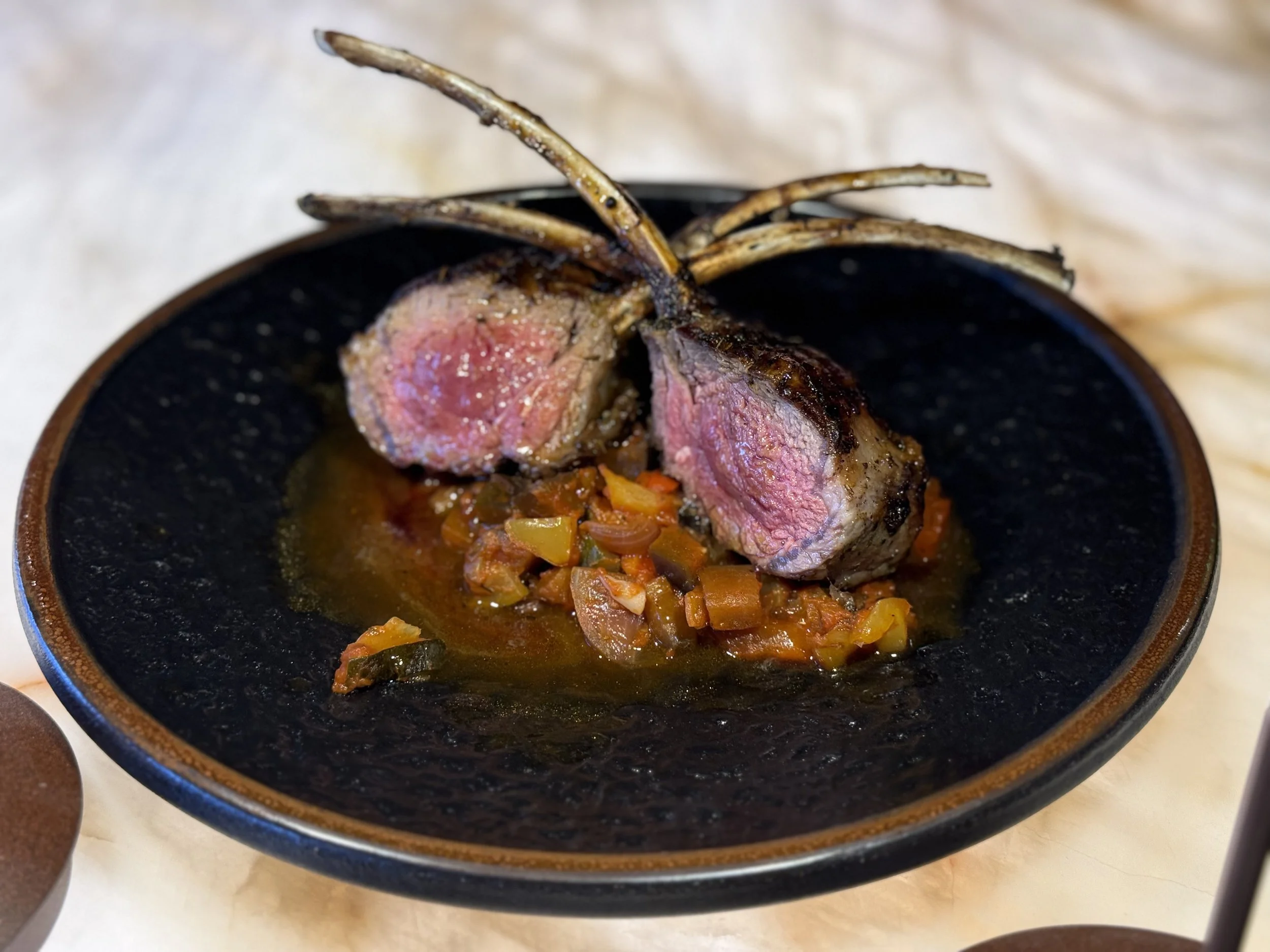 Two cooked lamb chops on a black plate with a side of stewed vegetables.
