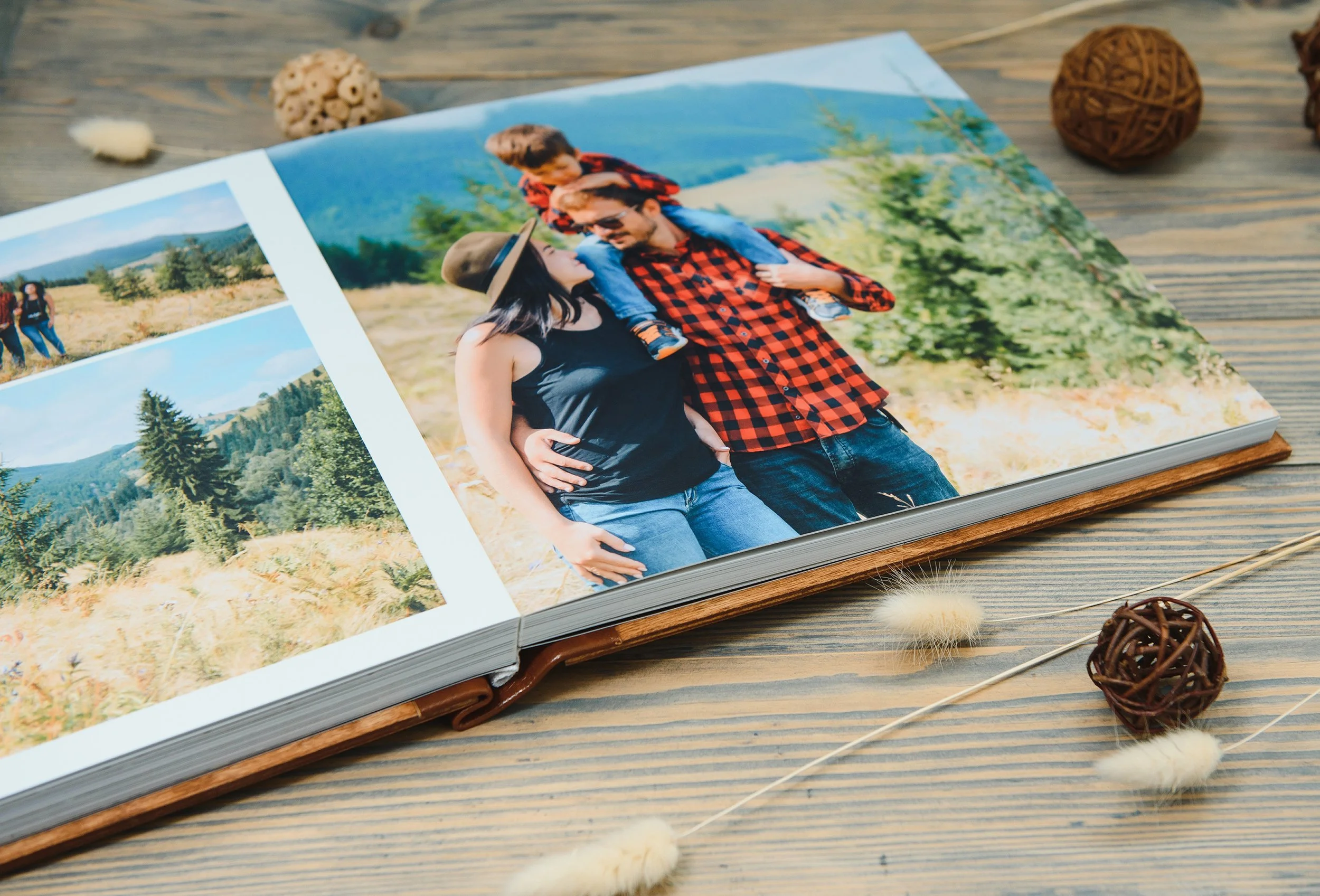 An open photo album on a wooden surface displaying a picture of a family of three outdoors in a natural setting, surrounded by decorative dried flowers and wicker balls.