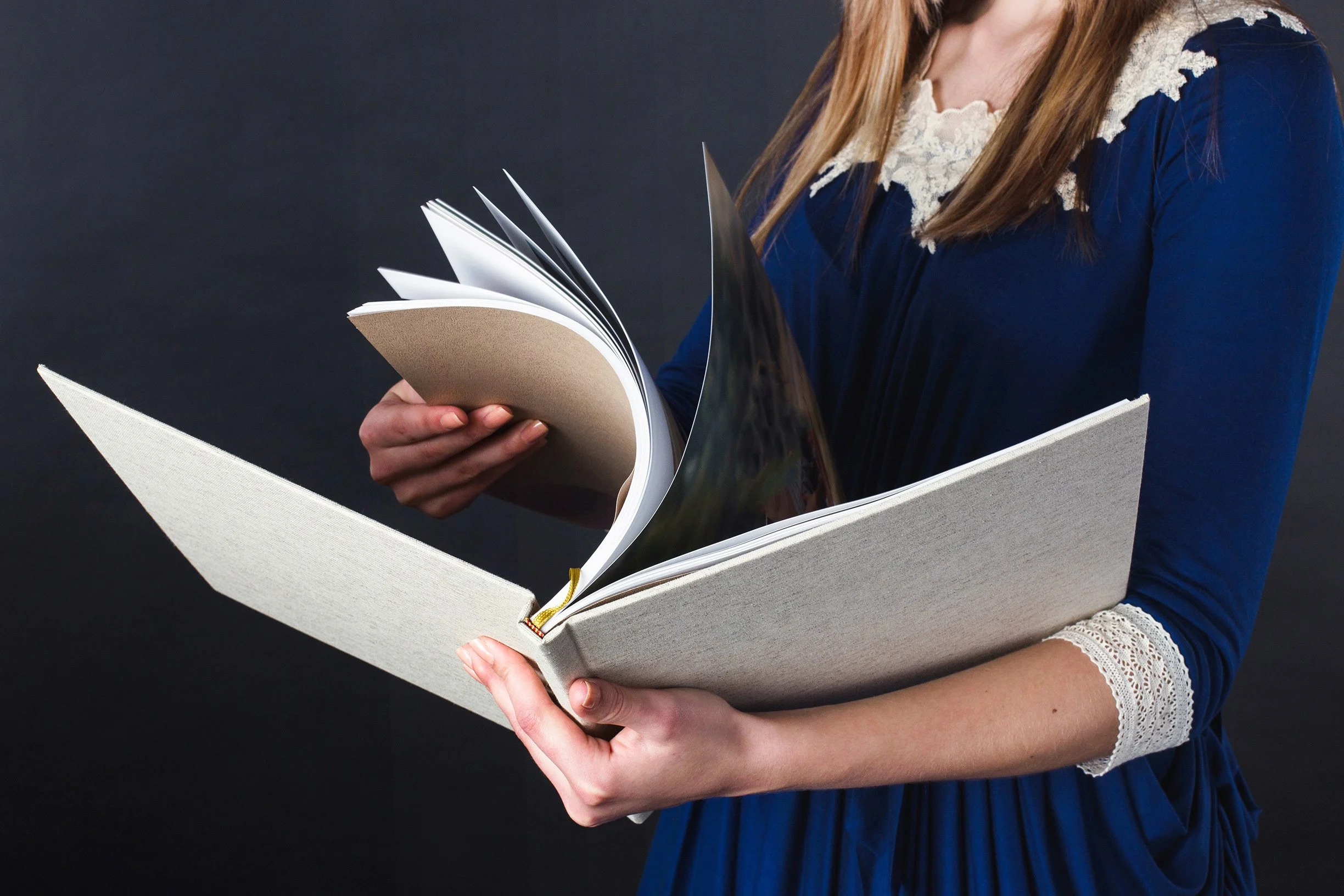 A woman wearing a blue dress with lace details on the collar and sleeves, holding an open large book and flipping through the pages against a dark background.