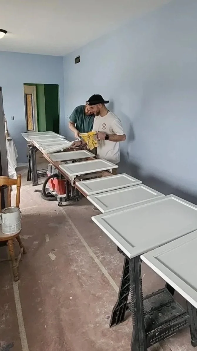 Two men are working on painting cabinet doors in a room with light blue walls, using paint and brushes.