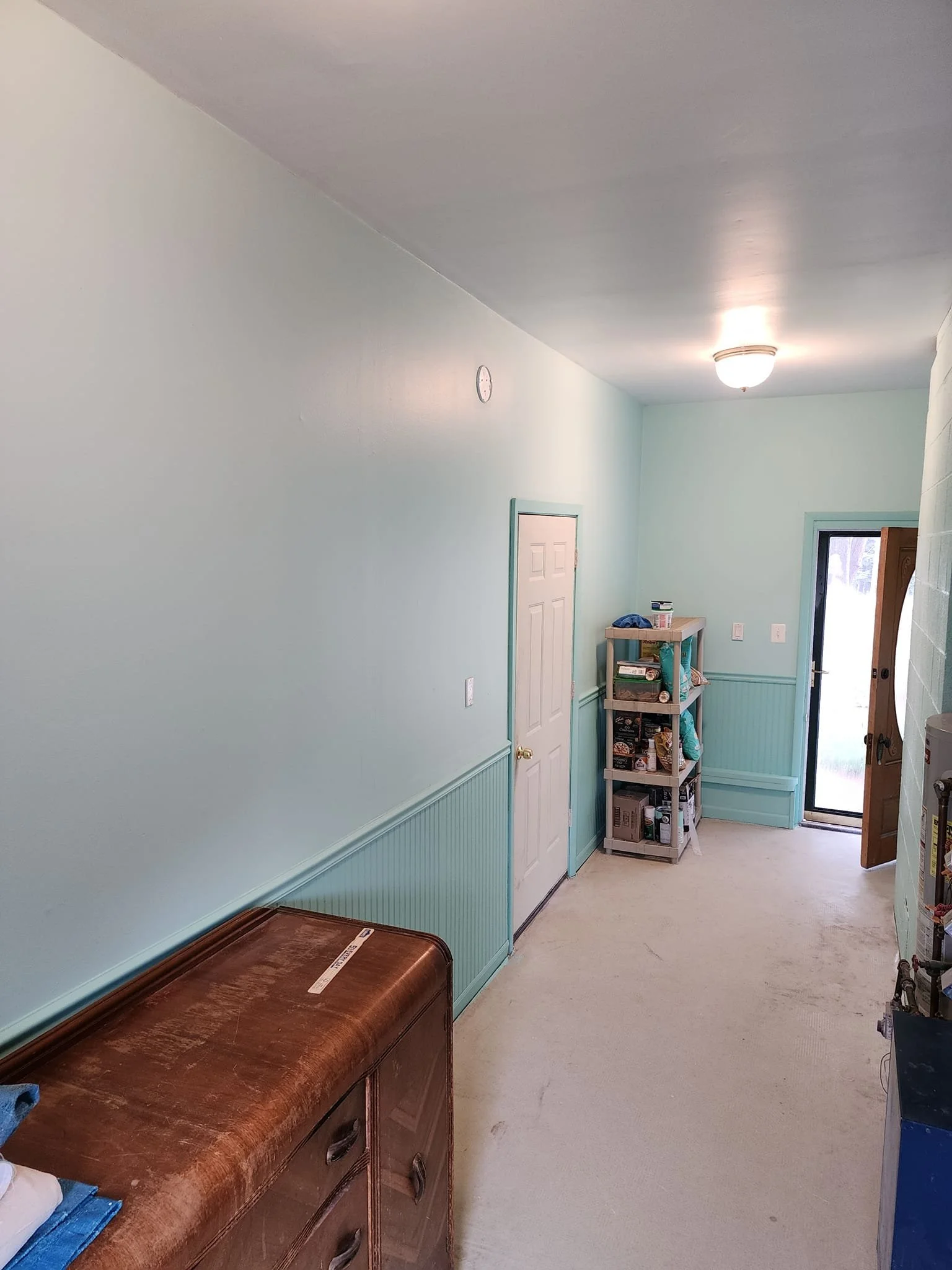 An interior hallway of a house with light green walls, a wooden cabinet on the left, a small shelving unit filled with various items near the door, and a glass door leading outside. The ceiling has a light fixture, and the floor is carpeted.