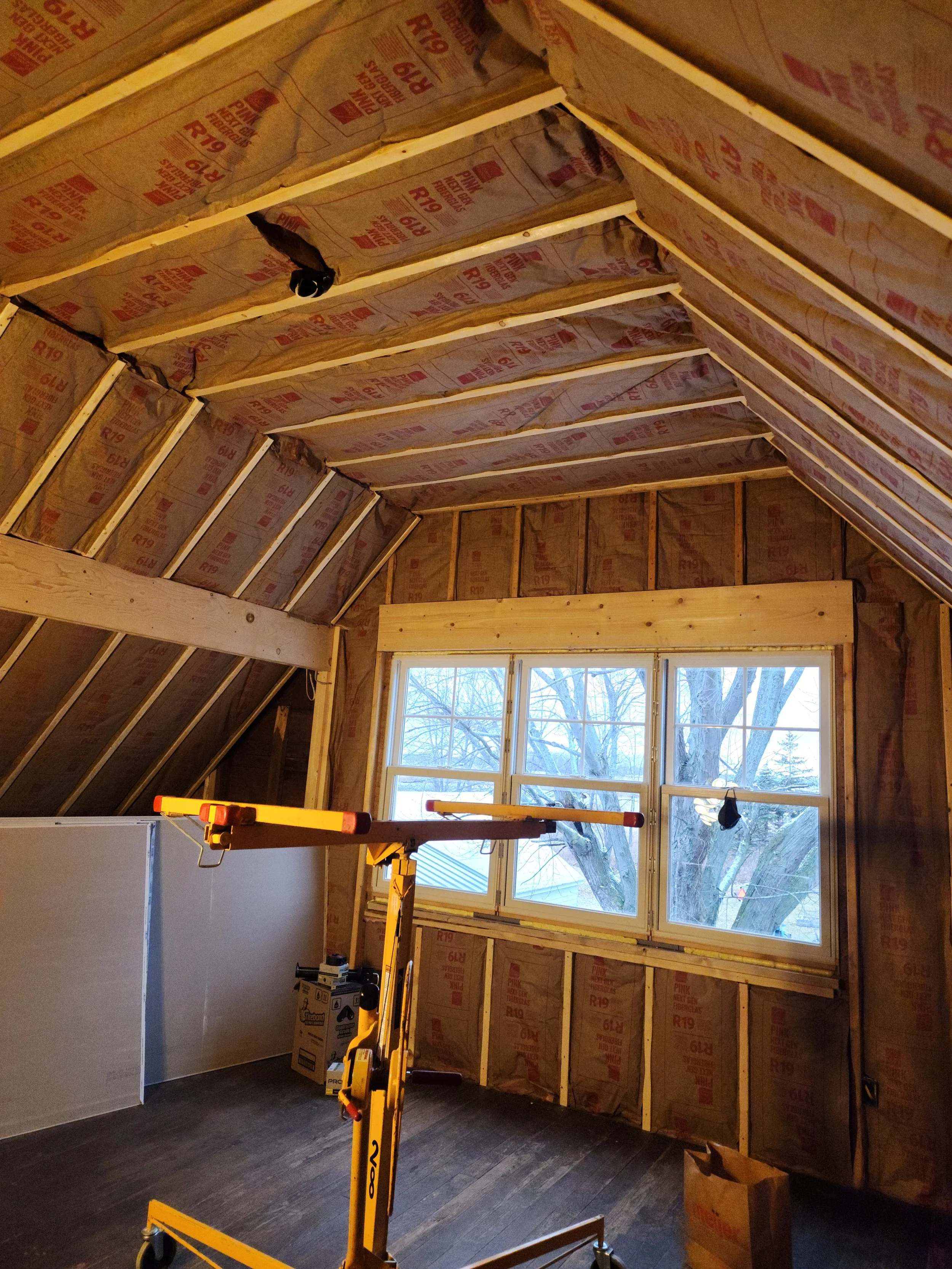 Interior of a room under construction with insulation on the walls and ceiling, a window showing trees outside, and construction equipment inside.