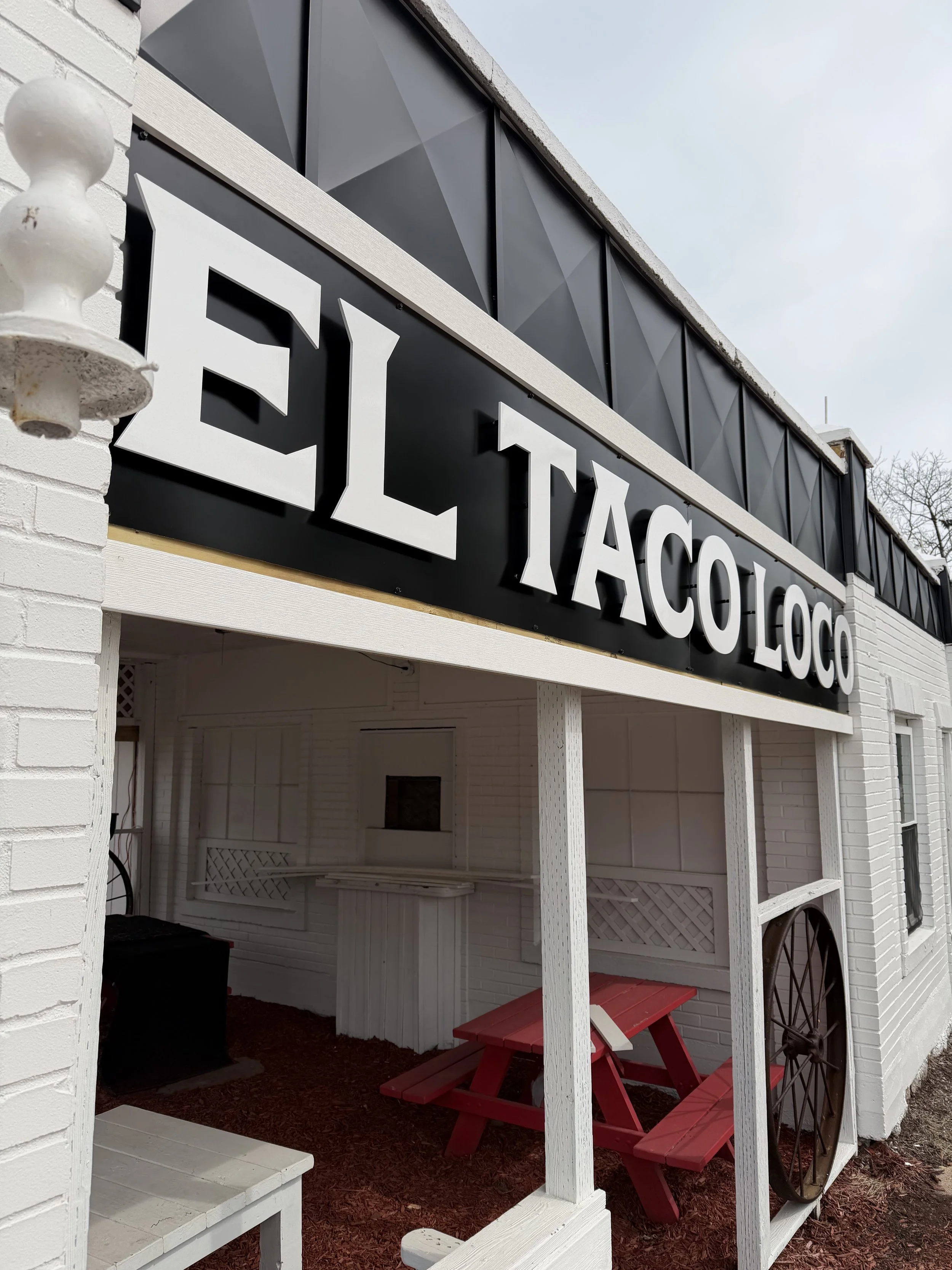 Exterior of a restaurant named EL TACO LOCO with white brick walls, a black sign with white raised letters, a red picnic table, a black wagon wheel, and a covered seating area.