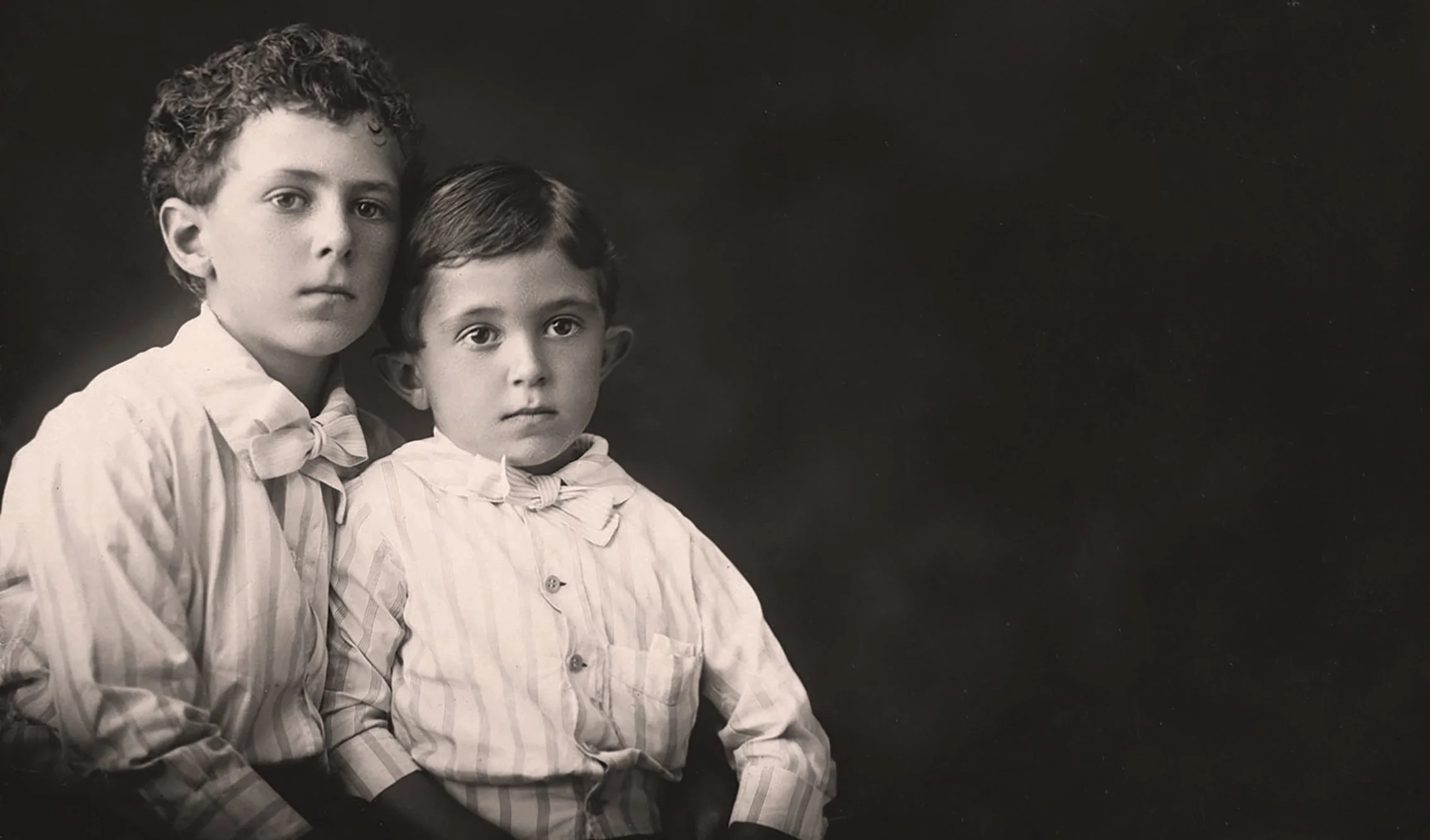 Black and white photograph of two young boys with serious expressions, dressed in striped pajamas, sitting against a dark background.