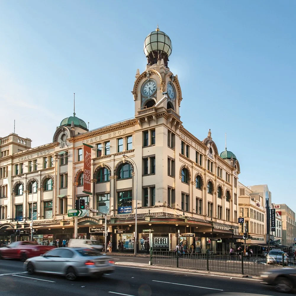 A historic building on a city street corner with a clock tower, featuring intricate architectural details, green domes, and a sign for Broadway.