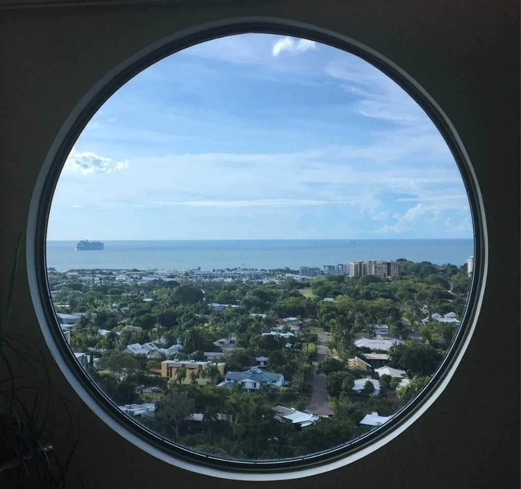 View of Darwin Harbour through a circular window showing a coastal cityscape with greenery, residential buildings, a distant ocean, and a cruise ship on the water.