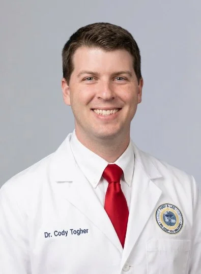 Professional portrait of a young male doctor in a white coat with a red tie, smiling against a light gray background.