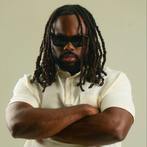 A man with dreadlocks wearing sunglasses and a light-colored polo shirt, crossing his arms and looking at the camera against a plain background.