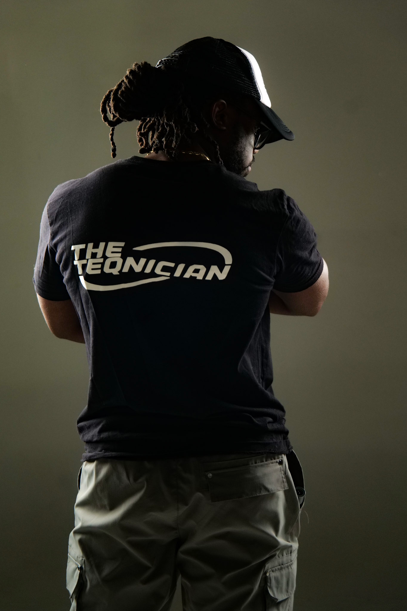A person wearing a black T-shirt with the text "The Technician" on the back, a black and white baseball cap, and beige cargo pants, standing against a plain background.