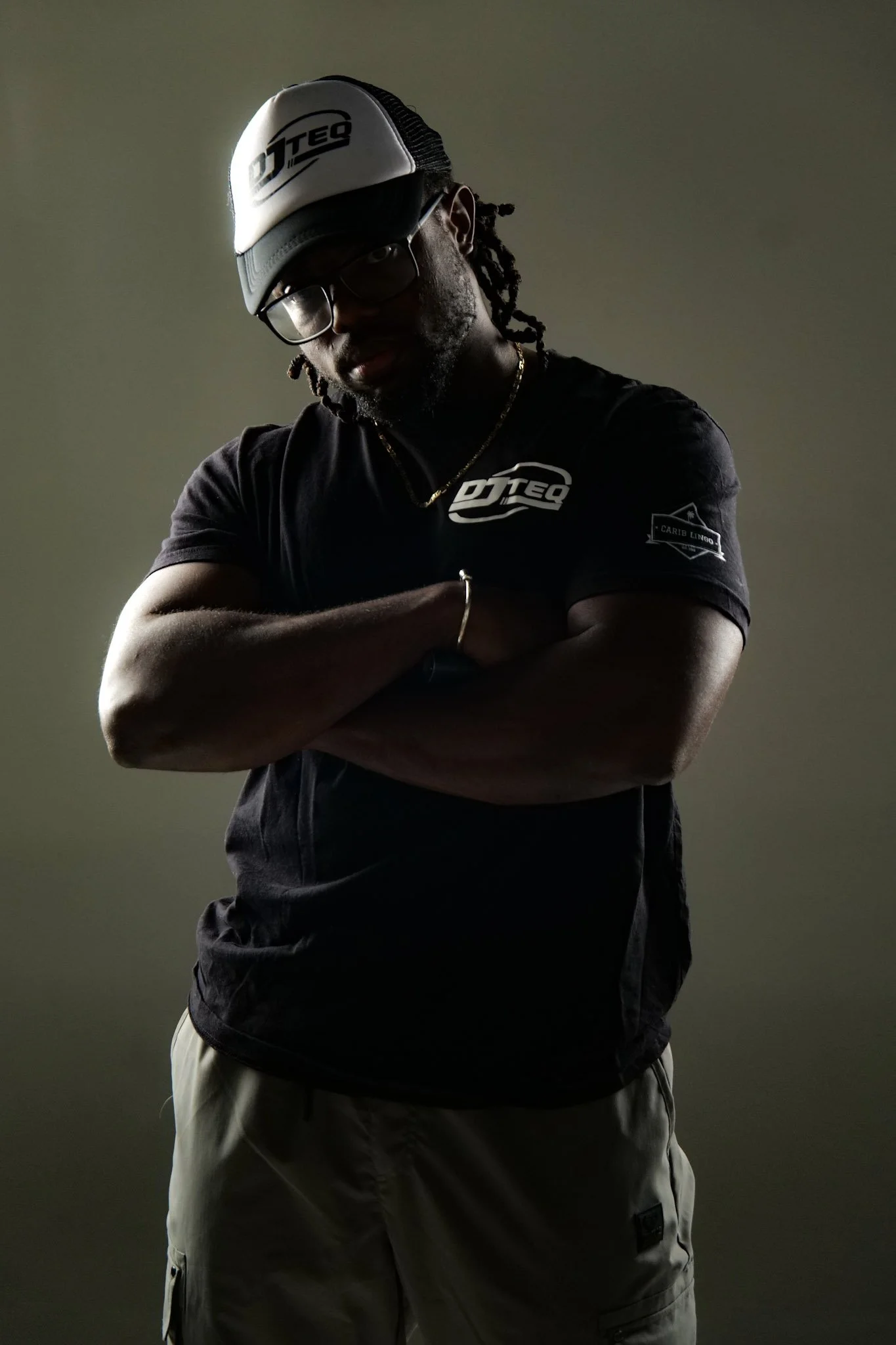 A man with glasses, wearing a black T-shirt, tan cargo shorts, a white and black cap, and a gold chain, stands with arms crossed against a plain background.