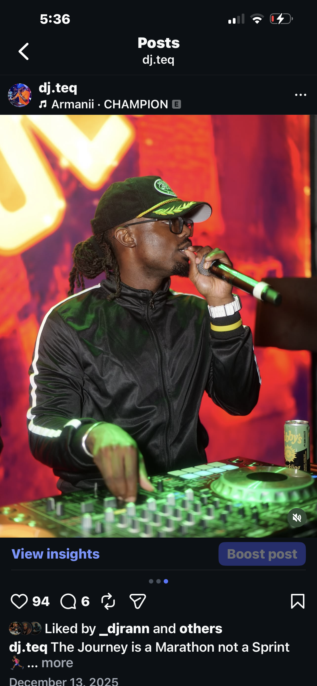 A DJ wearing a black tracksuit, glasses, and a black hat with a green logo, holding a microphone and operating a DJ controller with green lighting. The background is red and orange with blurred visuals.