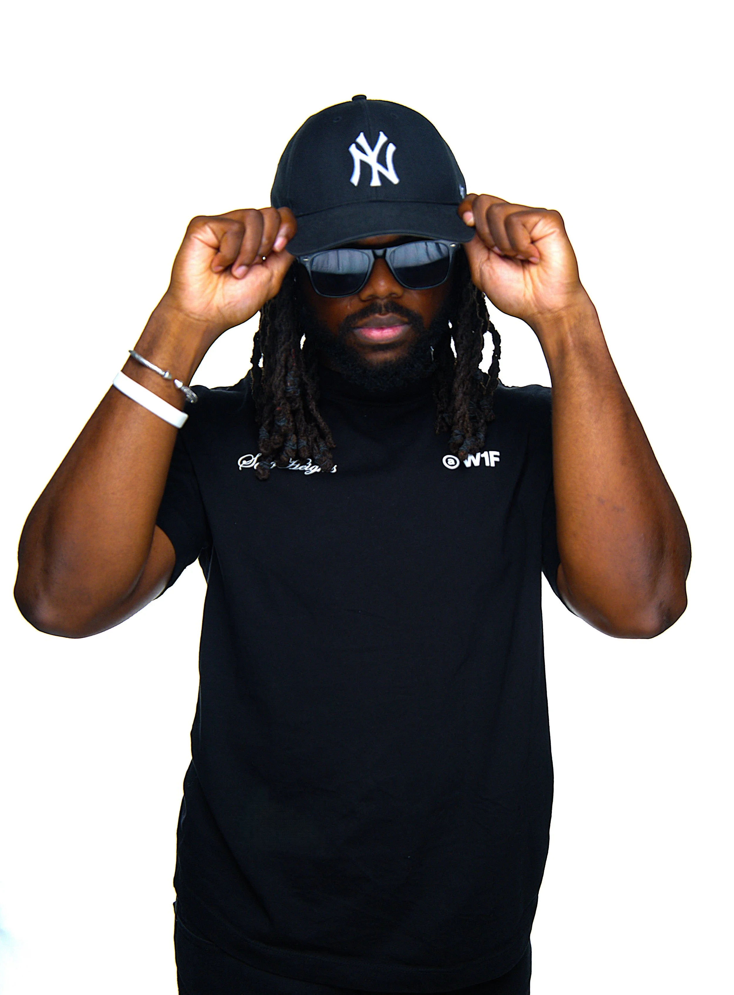 A man with dreadlocks wearing a black New York Yankees cap, sunglasses, and a black T-shirt with white embroidered text, adjusting his cap with both hands against a plain white background.