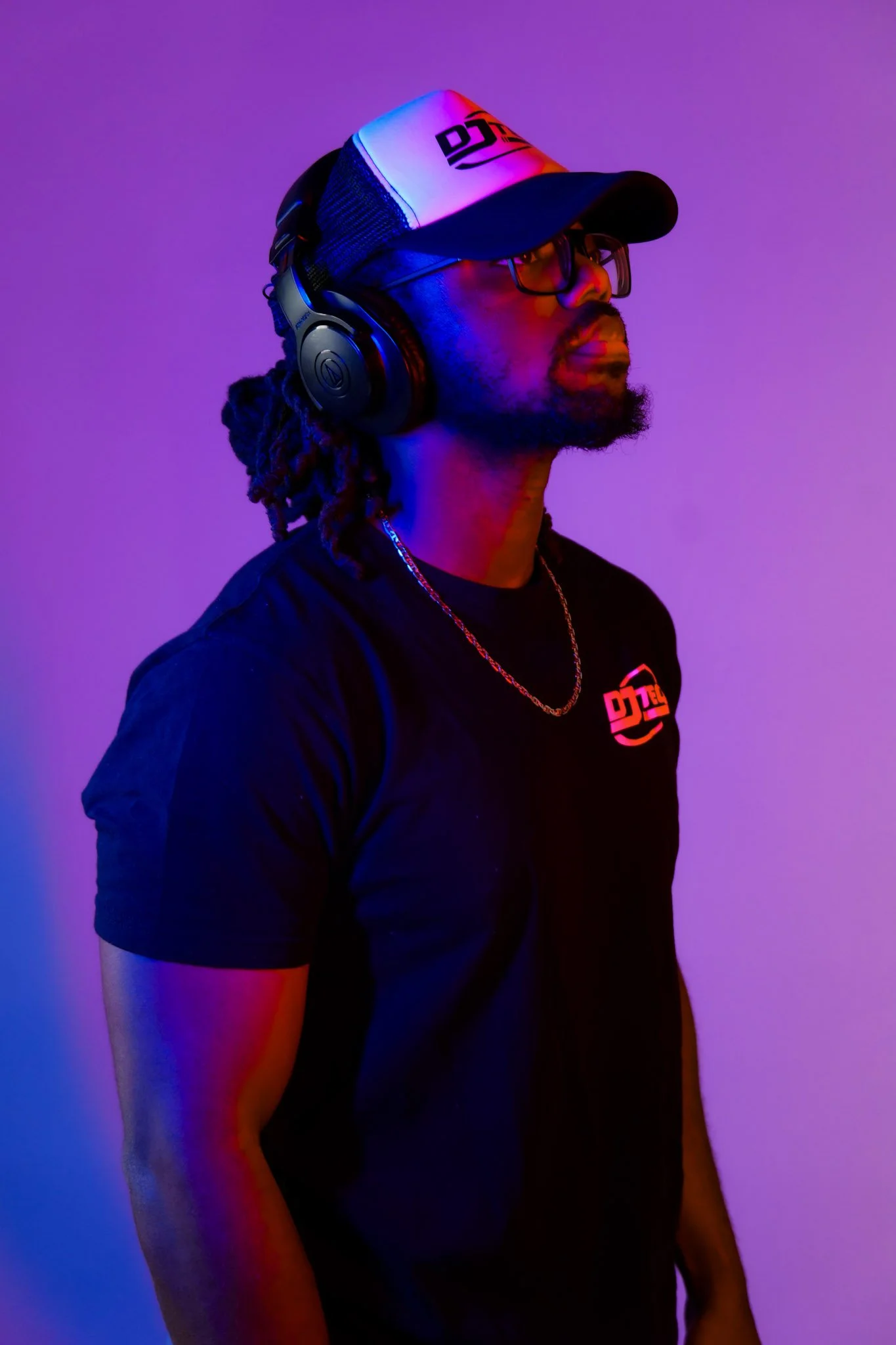 A man with dreadlocks wearing headphones and glasses, standing against a colorful purple and pink background.