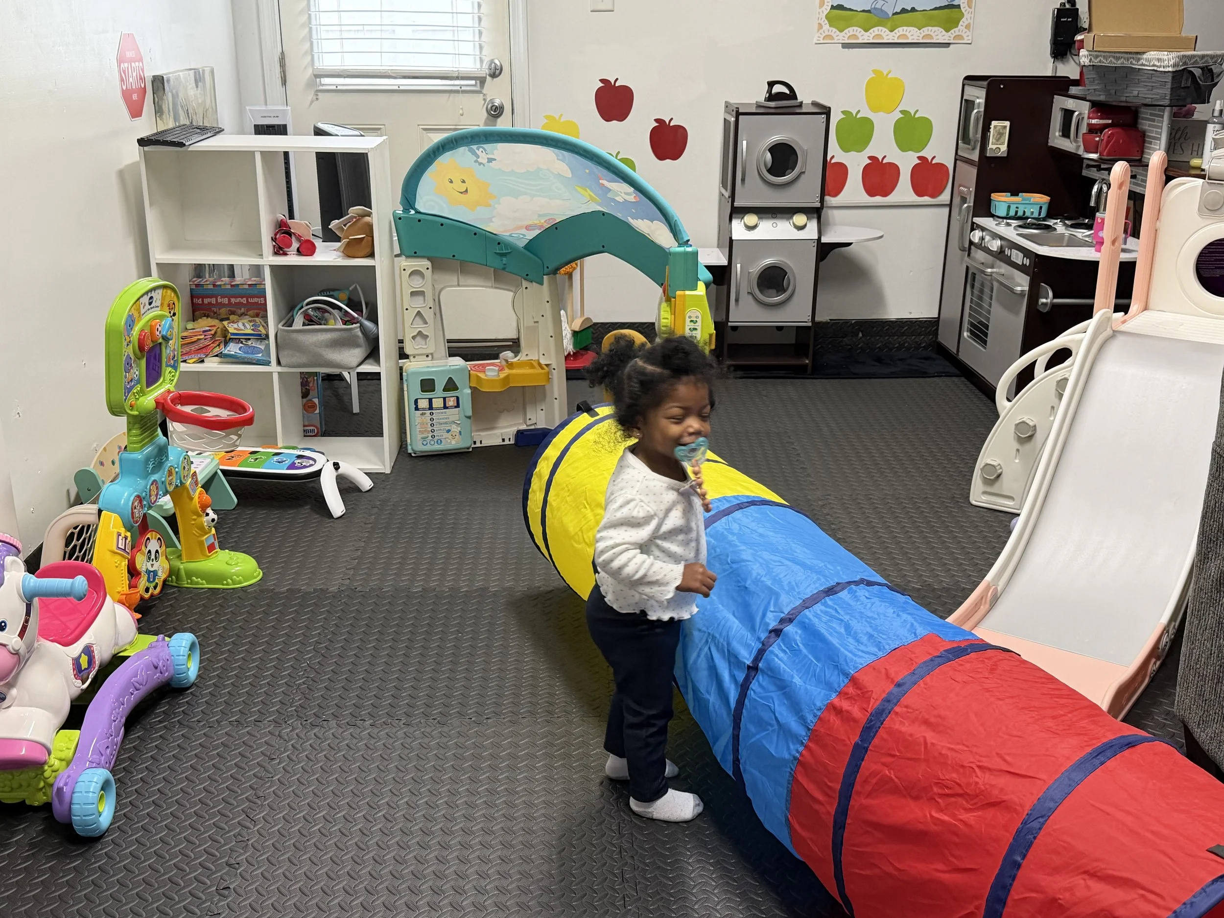 A young girl with a pacifier in her mouth playing in an indoor children's play area with toys, including a tunnel, slide, and a play kitchen, on a black rubber mat floor.