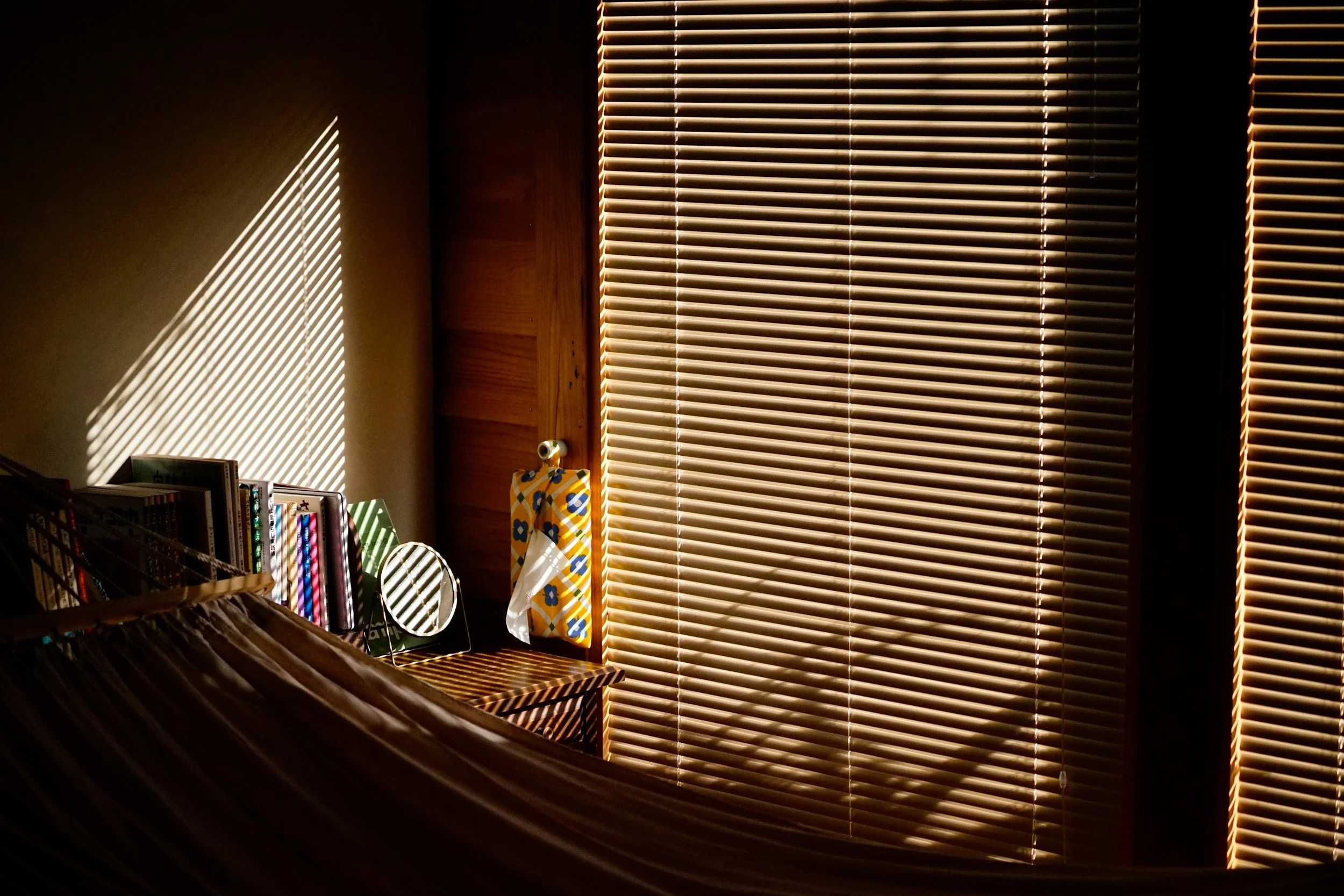 Sunlight streams through horizontal wooden blinds, casting striped shadows on a beige wall and objects on a bedside table, including books, a mirror, and decorative paper.