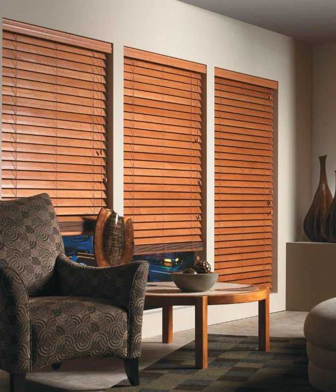 Living room with three large windows covered by closed wooden blinds, an upholstered armchair, and a wooden side table with decorative objects.