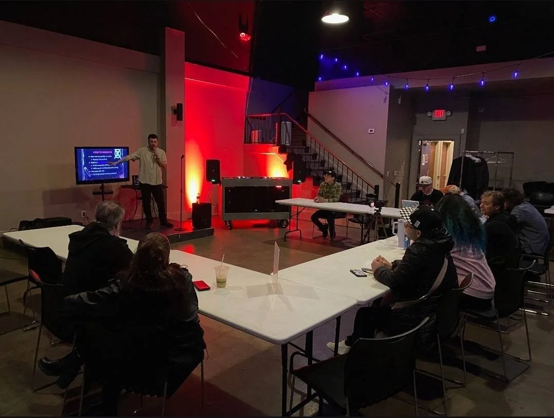 A man is giving a presentation to an audience seated at tables in a dimly lit room with red and blue lighting. There is a large display screen to his left showing a PowerPoint slide, and a microphone on his hand.