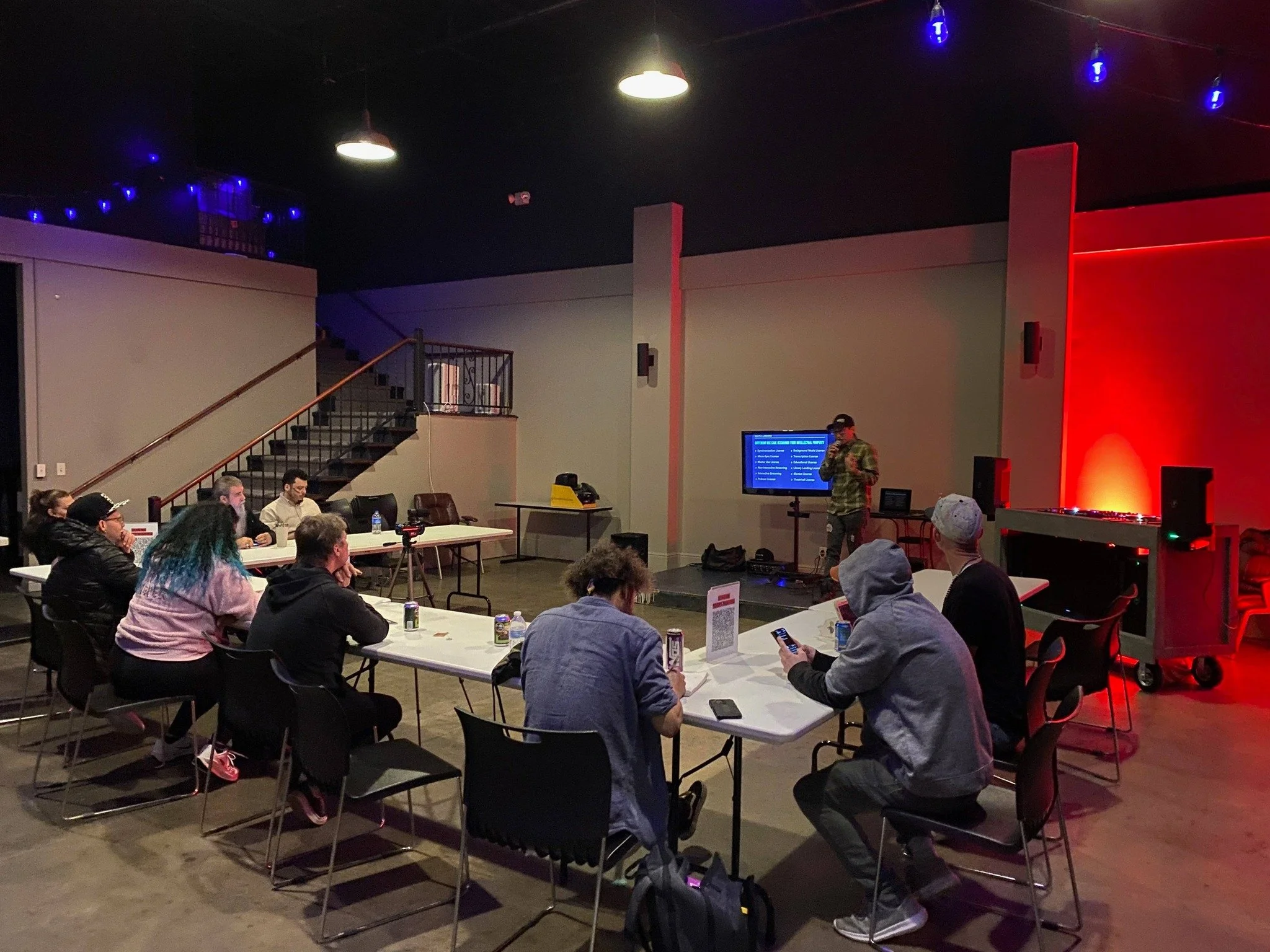 A group of people sitting at tables in a dark room with colorful lighting, attending a presentation or workshop with a speaker standing near a screen and a DJ setup in the background.