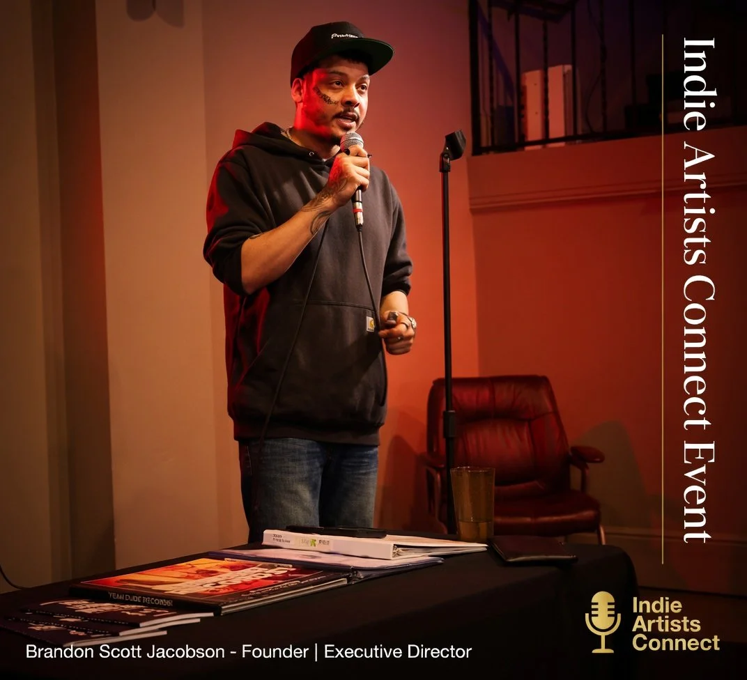 A man speaking into a microphone at an indoor event with a table of magazines and documents in front of him. He is wearing a black hoodie, a black cap, and has tattoos on his left arm. The event is called Indie Artists Connect, and the man identified