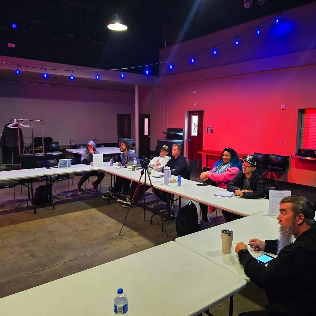A group of people sitting at a long white table in a dimly lit room with colorful lighting, some looking at their phones or talking, with a coat rack, water bottles, and notebooks on the table.