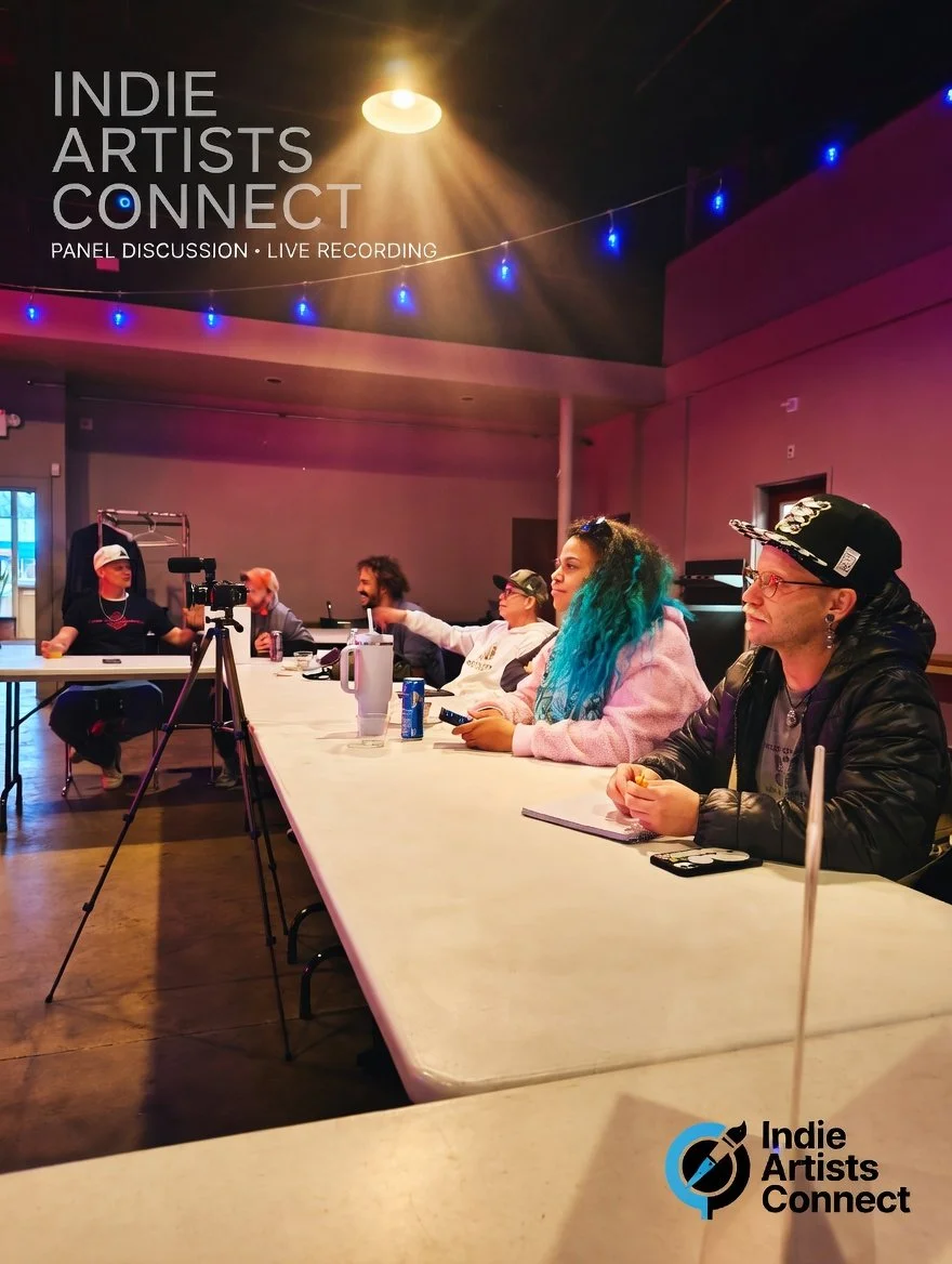Group of people sitting at a long table during a panel discussion titled "Indie Artists Connect" with a camera set up for recording, and blue string lights hanging overhead, in a venue with purple and pink lighting.