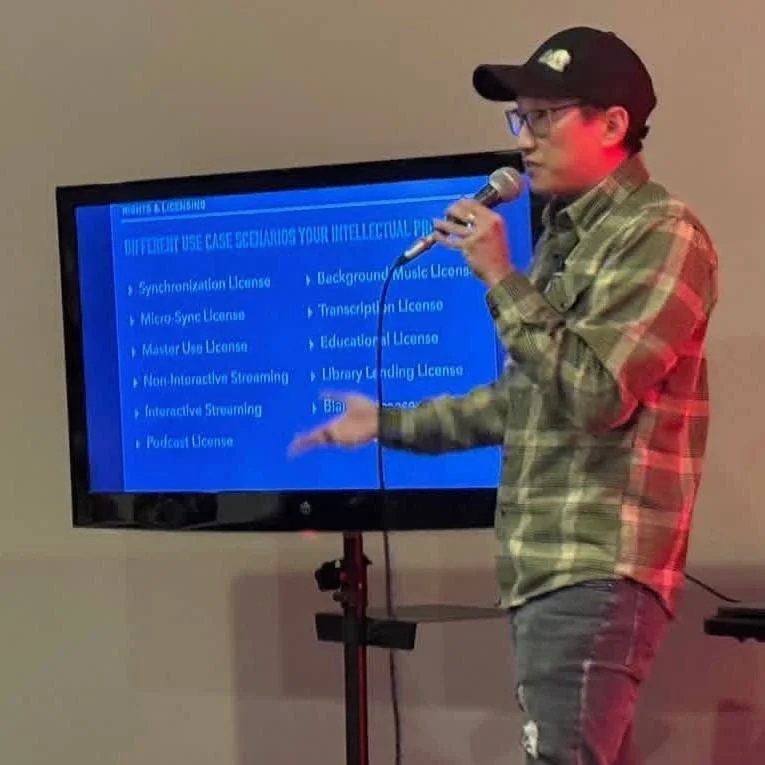 A man wearing glasses, a black cap, and a plaid shirt giving a presentation with a microphone in hand, pointing at a slide on a blue screen with white text.