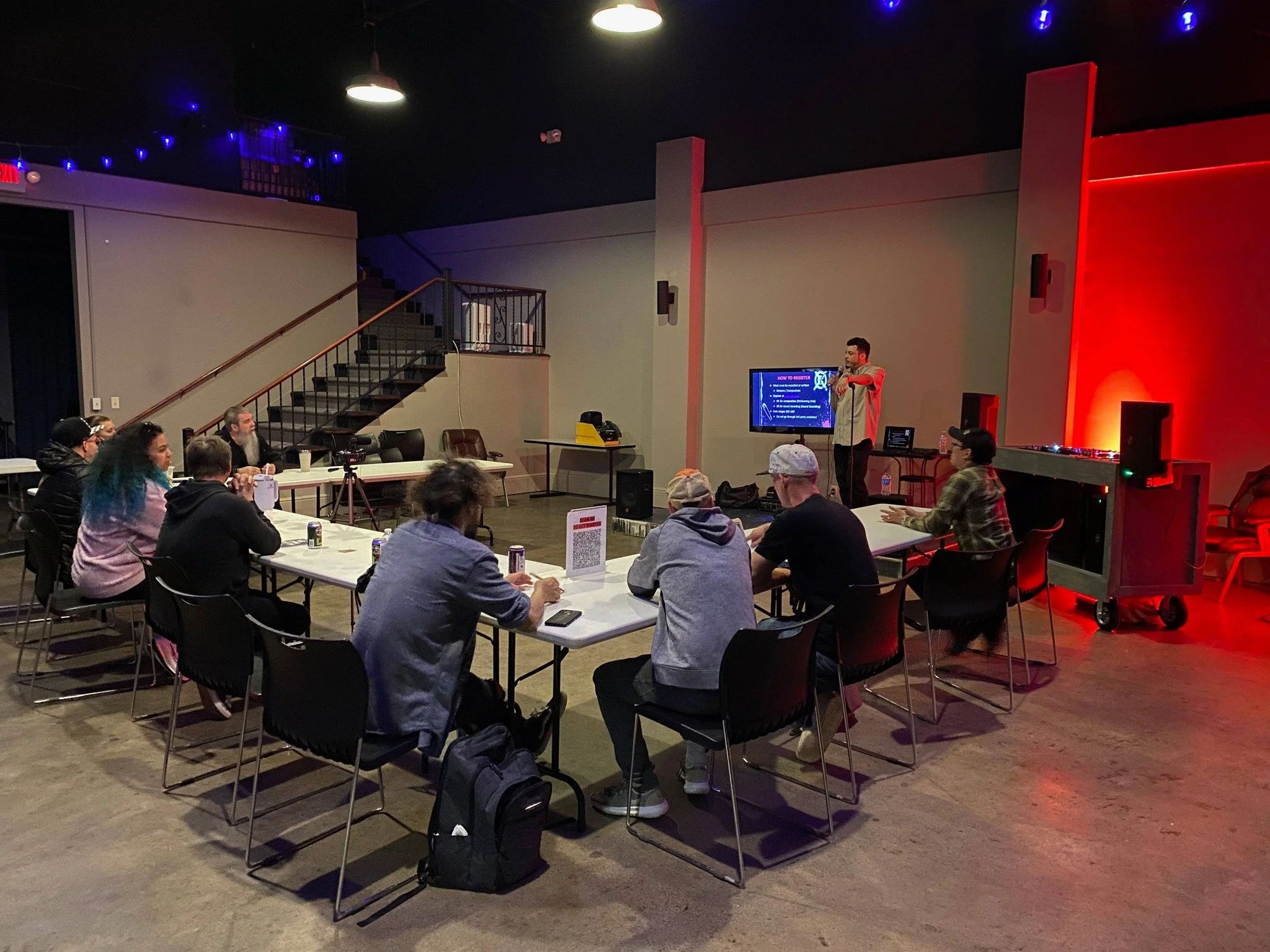 A group of people seated at tables attending a presentation in a dimly lit room with a speaker standing near a screen. The room has colorful lighting and a staircase leading to an upper level.