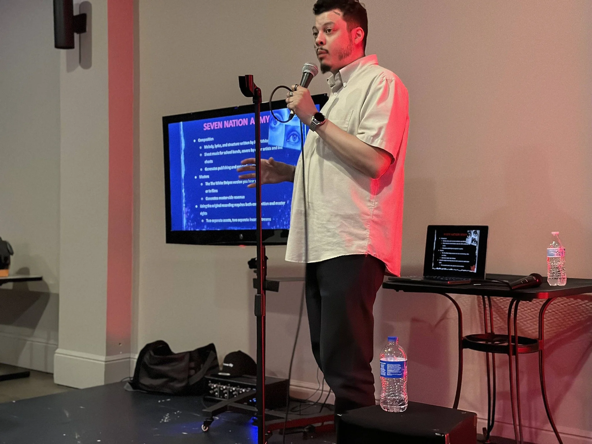 A man is standing on a stage giving a presentation, holding a microphone in his right hand and gesturing with his left. He is wearing a light-colored short-sleeved shirt, black pants, and a watch. Behind him, a large screen displays a slide with a bl