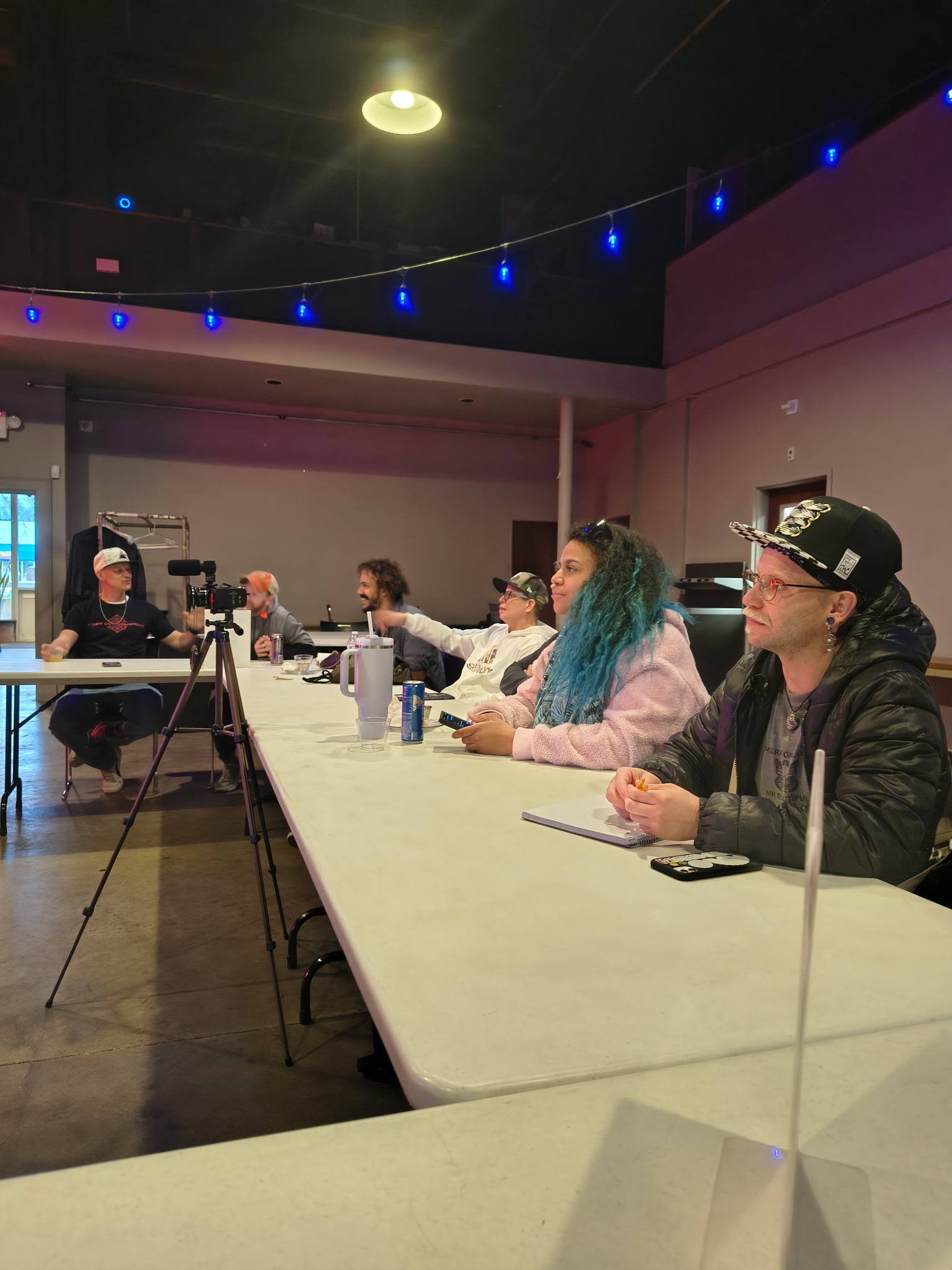 People sitting at a long table in a meeting or workshop room, with a camera on a tripod facing them. The room has purple and blue lighting, and some are taking notes or using their phones.