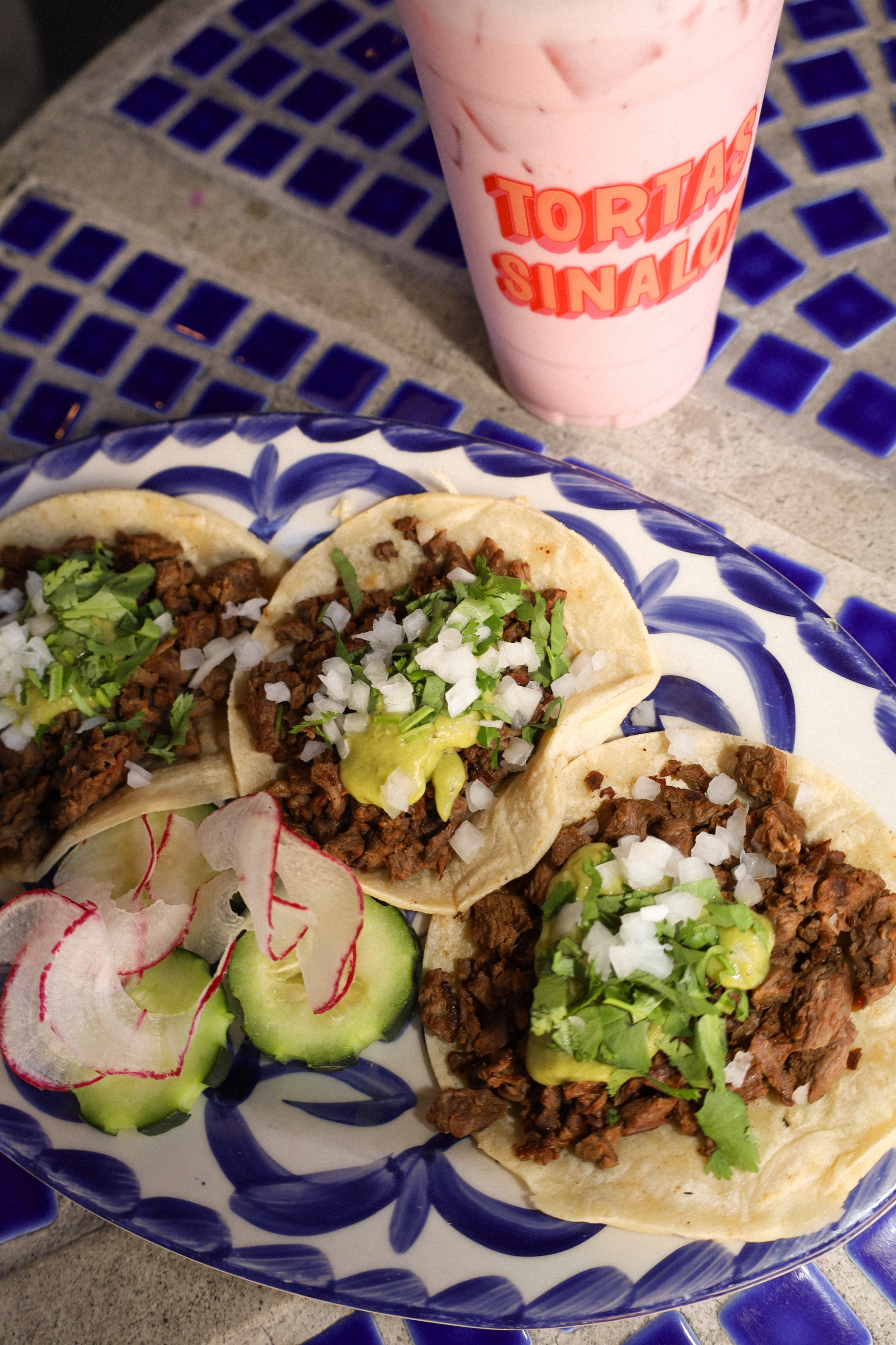 A plate with three tacos topped with chopped onions, cilantro, and avocado, along with cucumber and radish slices, on a blue and white patterned plate. A pink milkshake with the words "TORTAS SINALOA" written in red is in the background.