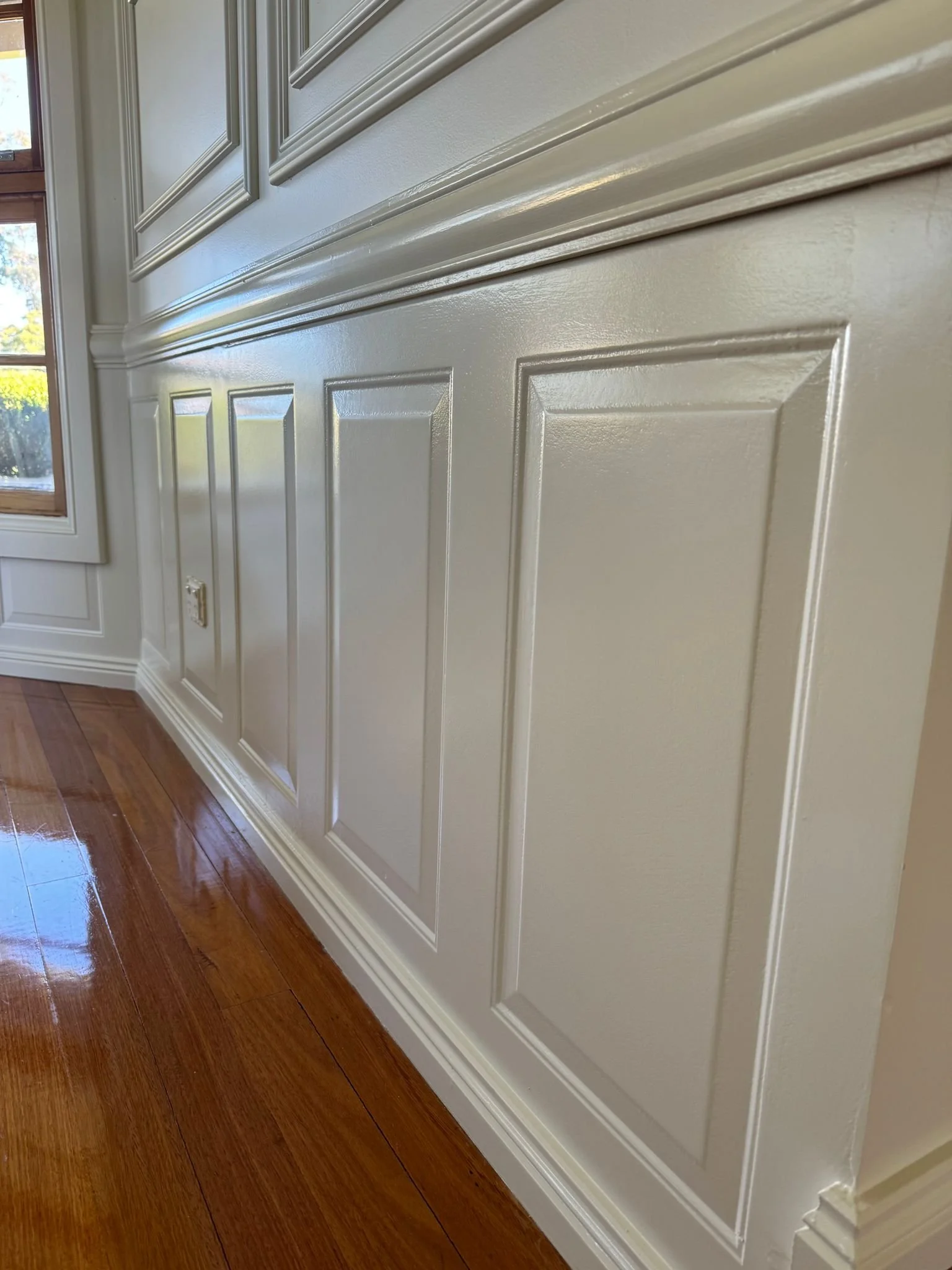 Close-up of a white painted wainscot wall panel with decorative molding, next to a window and wooden floor.