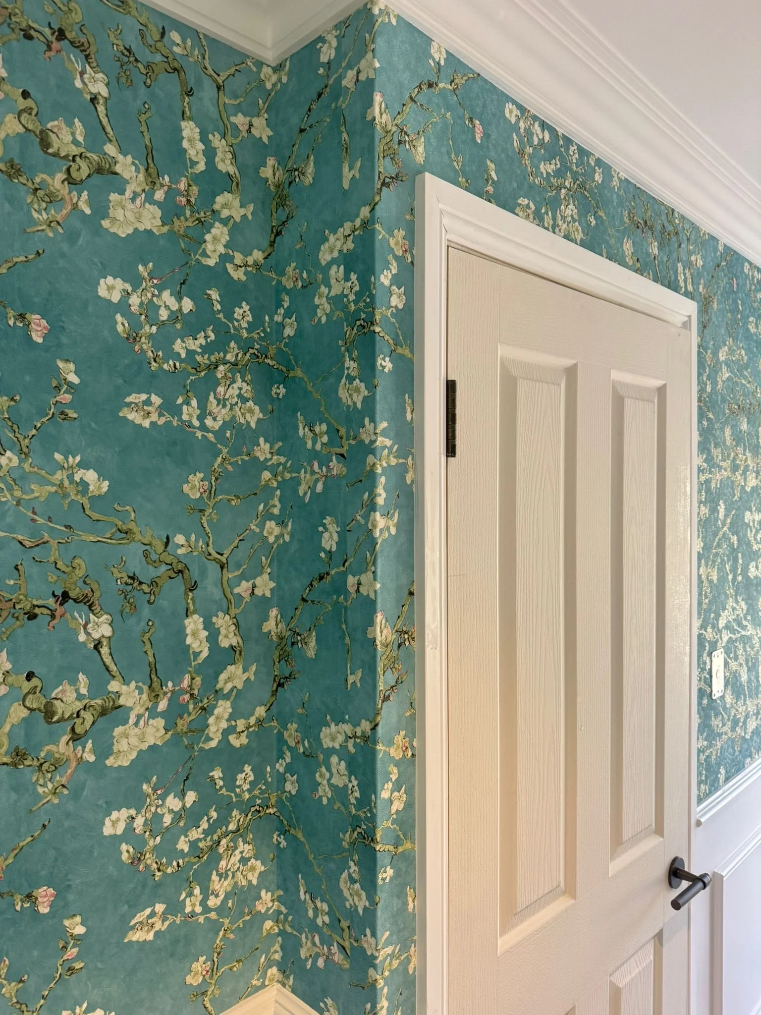 Close-up of a corner of a room with blue floral wallpaper featuring branches and white blossoms, next to a white paneled door with a black handle.