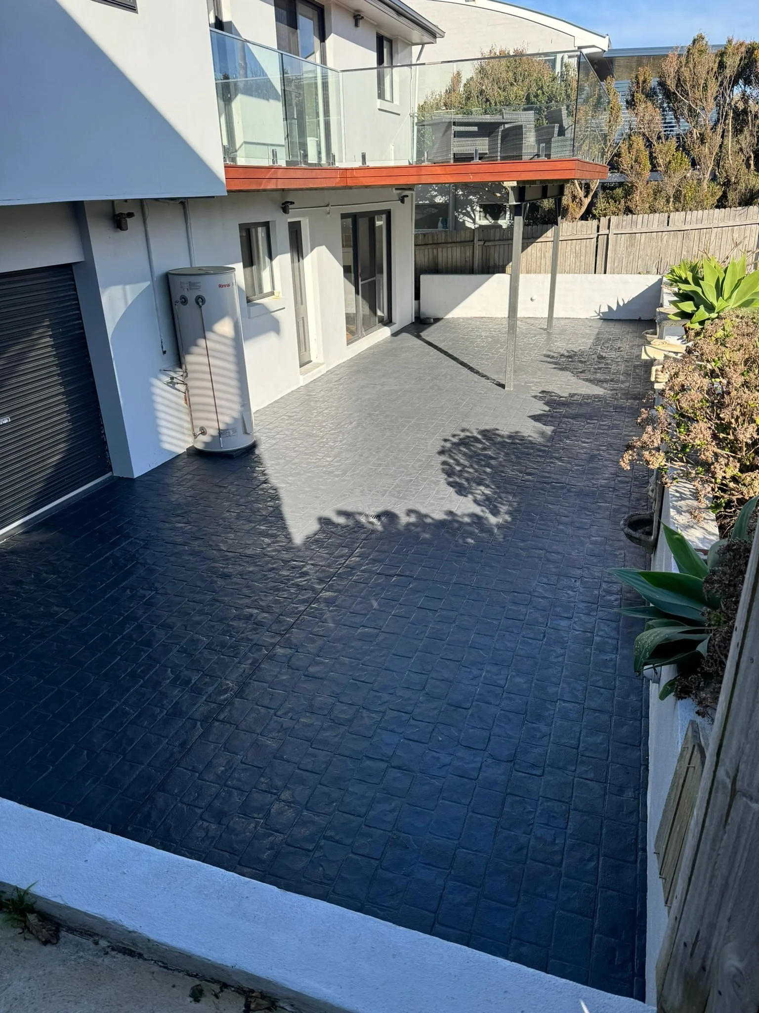 Modern backyard patio with black tiled floor, glass railing balcony, and some outdoor furniture in the background. There is a water heater on the left side near the house wall, and a garden with plants on the right side.