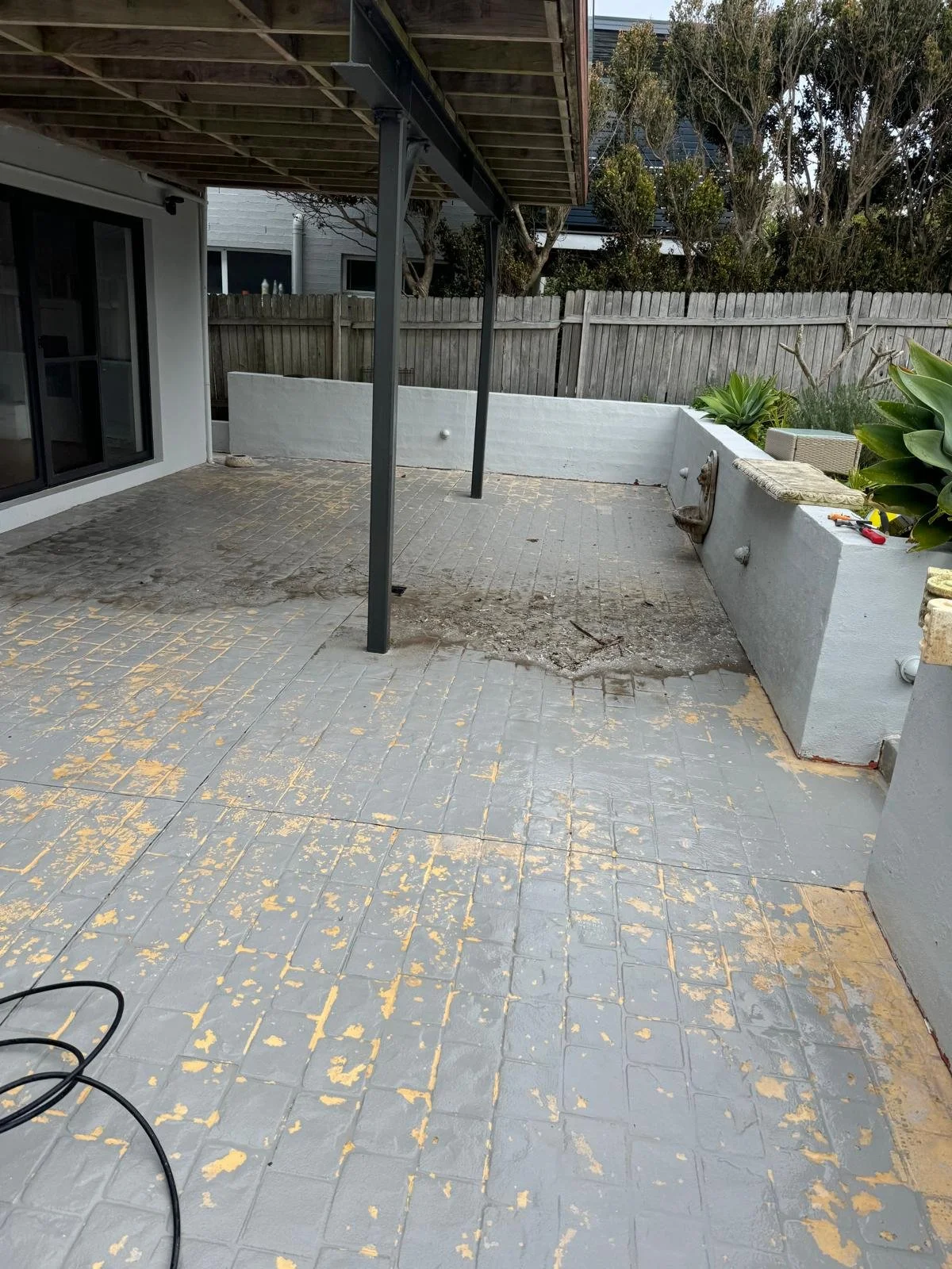 A backyard patio area with a tiled floor, partially stripped of old paint or coating, and some tools and materials on the edges. A white painted retaining wall, greenery, and a wooden fence are in the background.