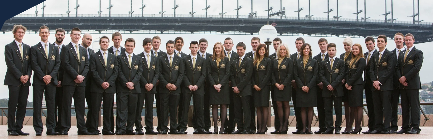 A group of young adults in formal black suits with gold accents, standing outdoors in front of a bridge and river, posing for a group photo.